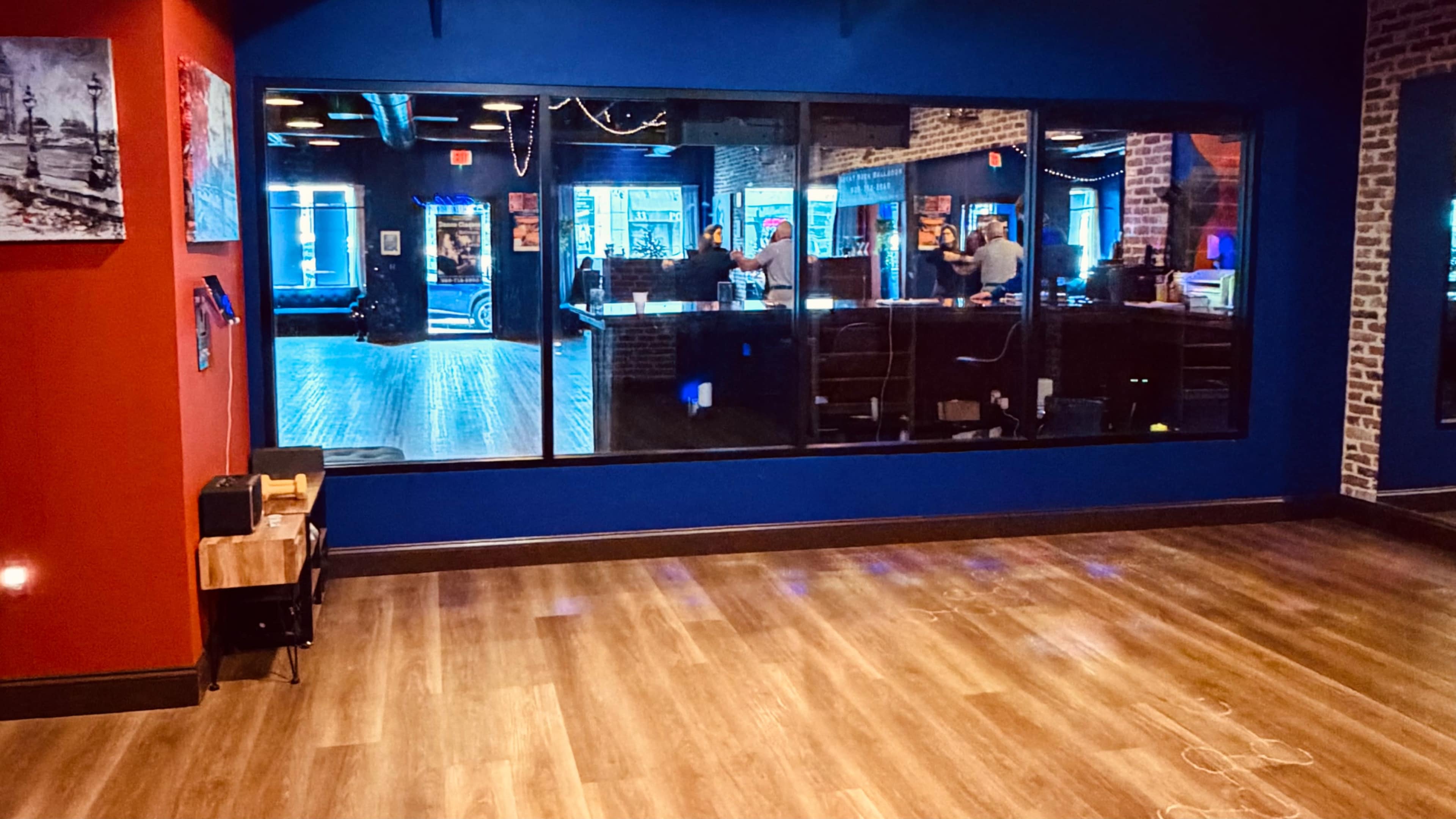 The image shows a large blue wall with a window that overlooks a brightly lit interior space featuring wooden flooring and a bar area.