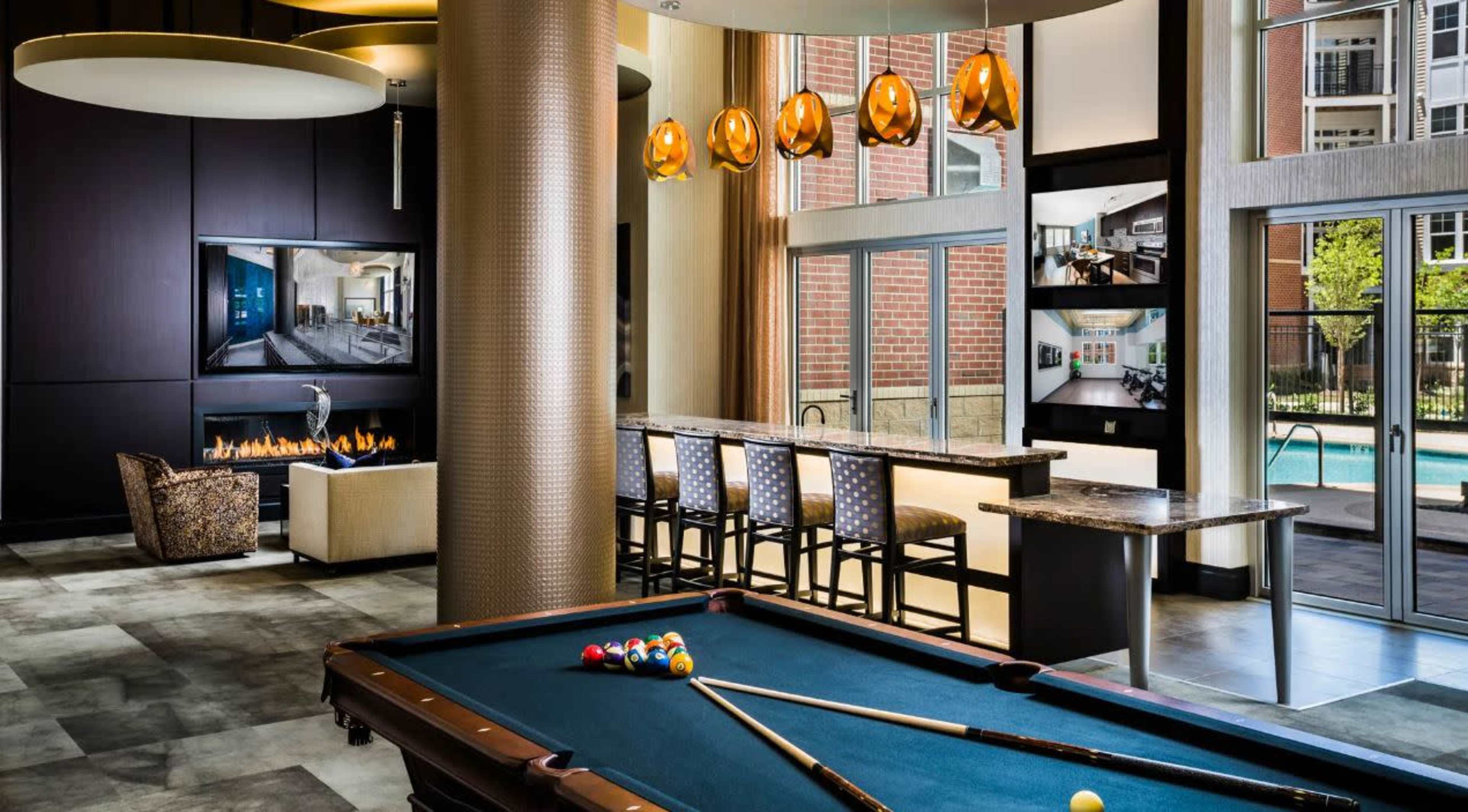 A modern lounge area with a pool table, a bar with stools, and large windows overlooking an outdoor pool.