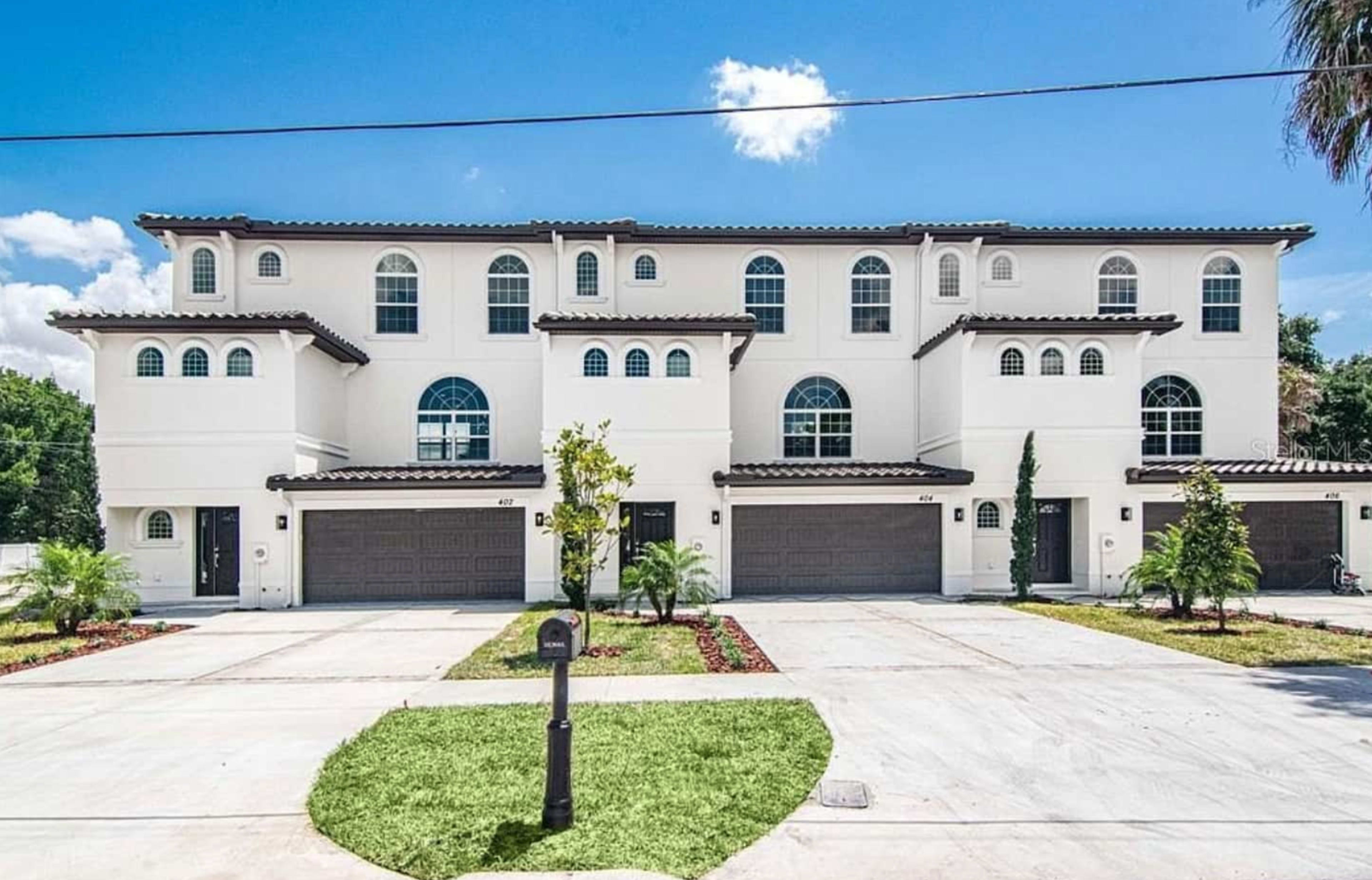 The image shows a row of three modern white multi-family homes with arched windows and a paved driveway, all set against a clear blue sky.