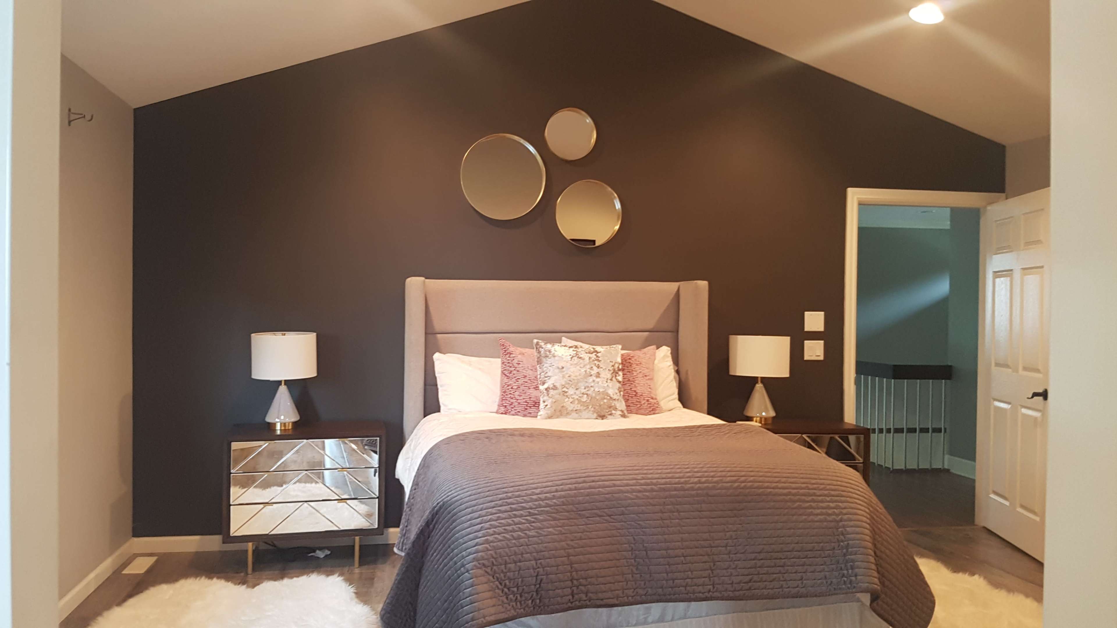A modern bedroom features a gray upholstered bed with decorative pillows, two mirrored nightstands, and three circular mirrors on a dark accent wall.