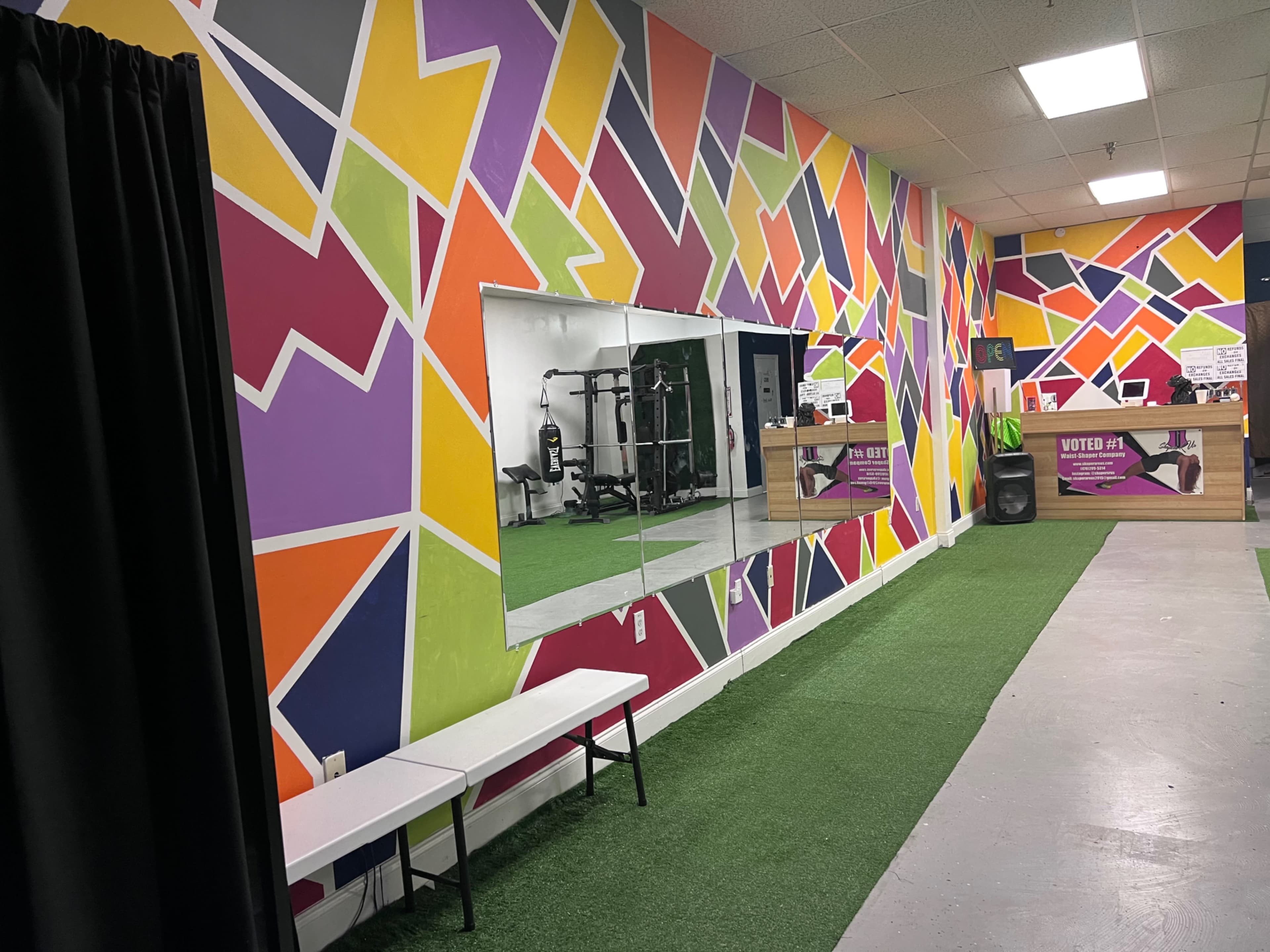 A brightly colored gym interior with a geometric mural on the walls, a mirror, a bench, and a patch of green turf.