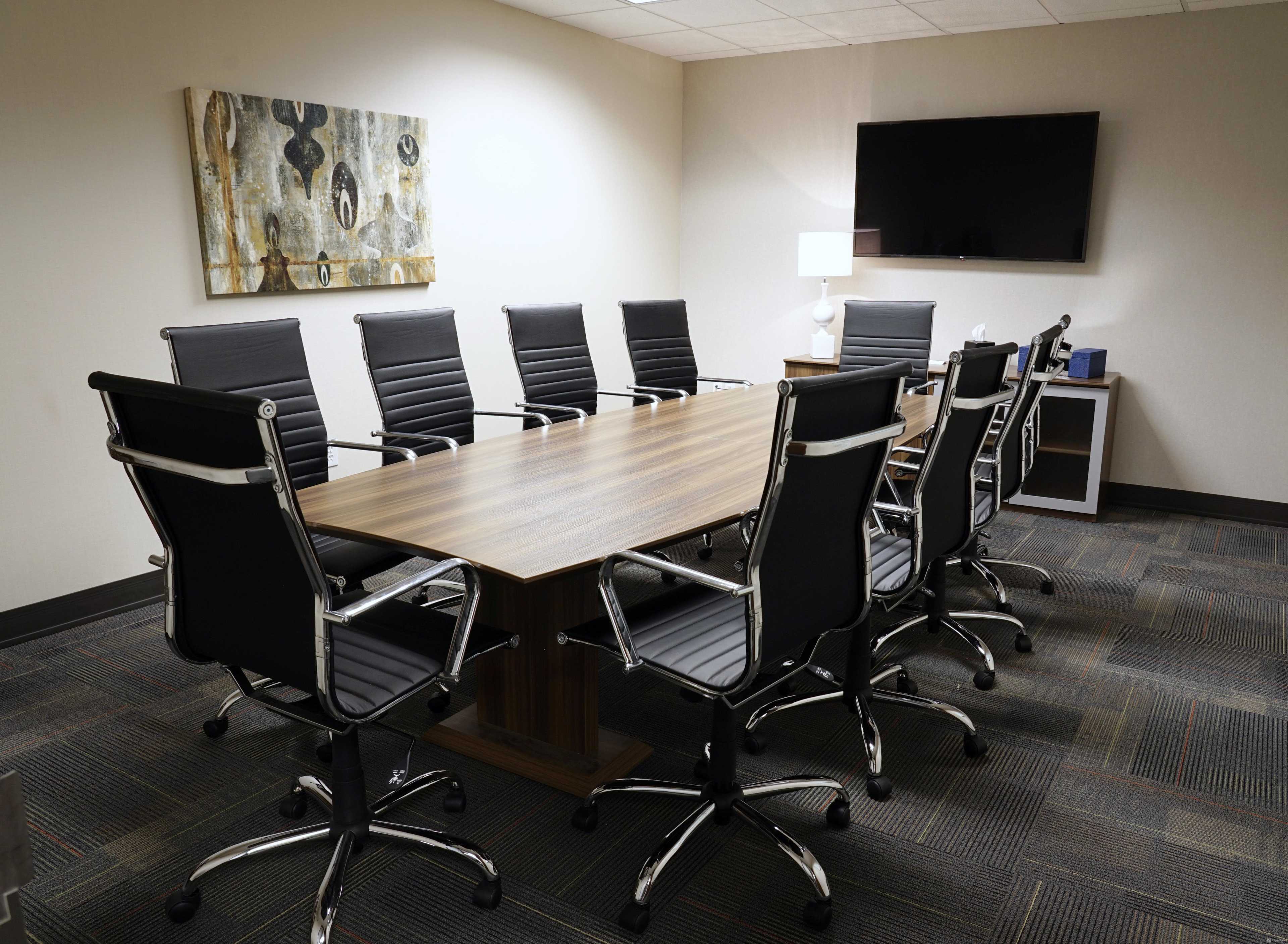 A conference room with a long wooden table surrounded by eight black office chairs, a television mounted on the wall, and a piece of art hung above the table.