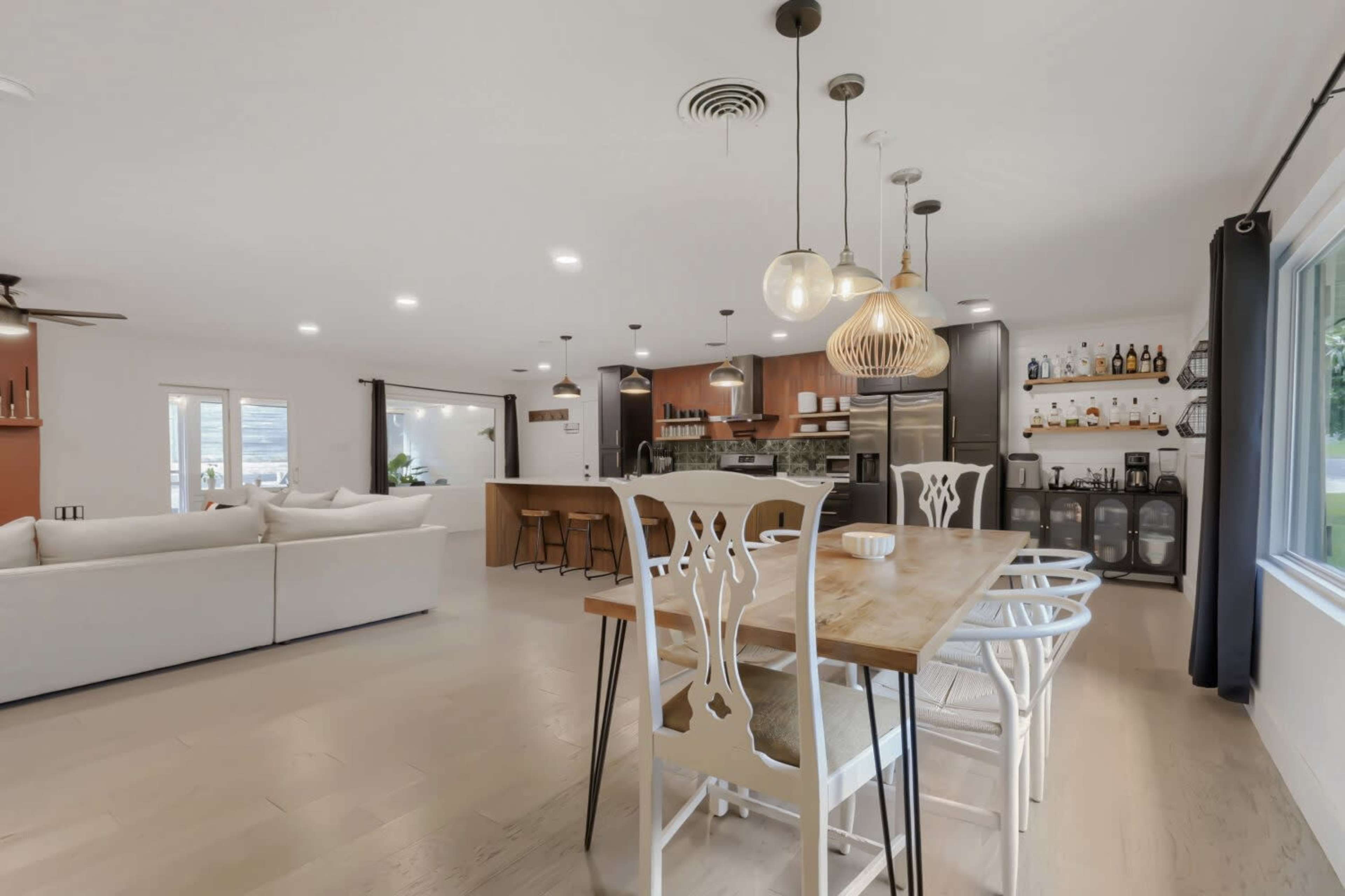 The image shows a modern dining area with a wooden table and white chairs adjacent to an open kitchen featuring a countertop and bar stools.