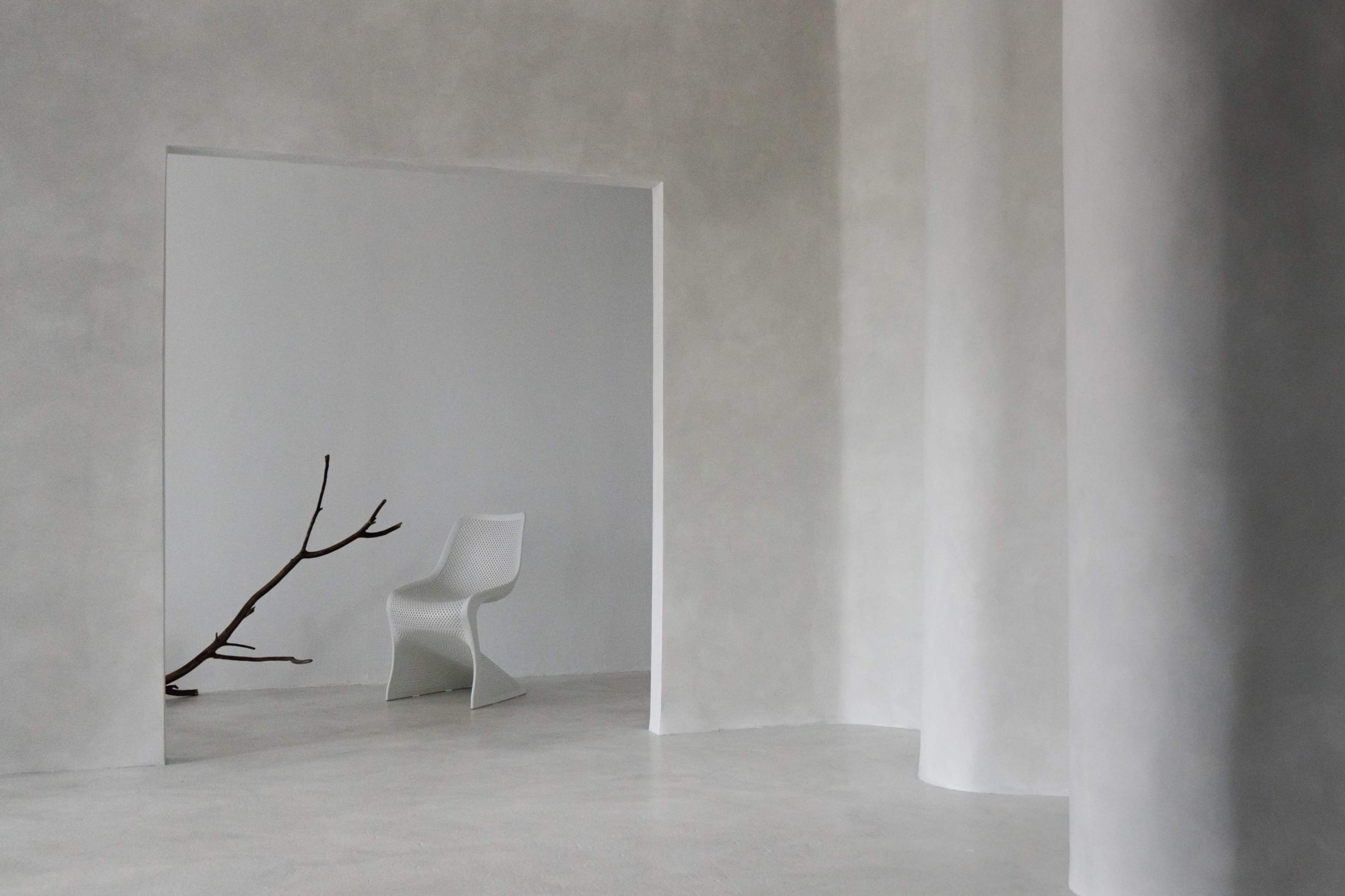 The image shows a minimalist interior space featuring a white chair and a bare branch positioned against a smooth, light-colored wall.