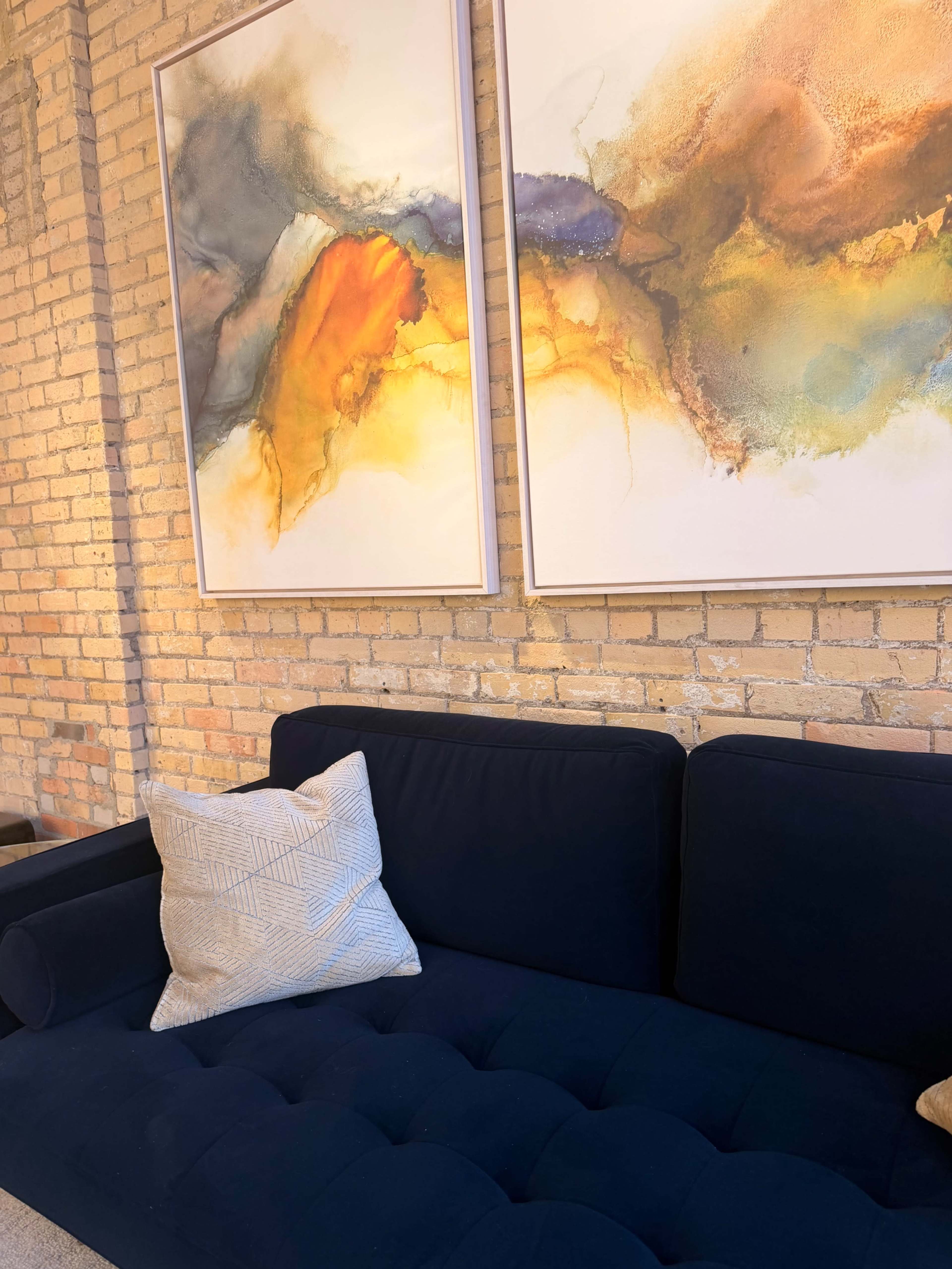 The image shows a navy blue couch with a patterned throw pillow, positioned against a wall adorned with two large abstract paintings in warm tones, alongside exposed brick.