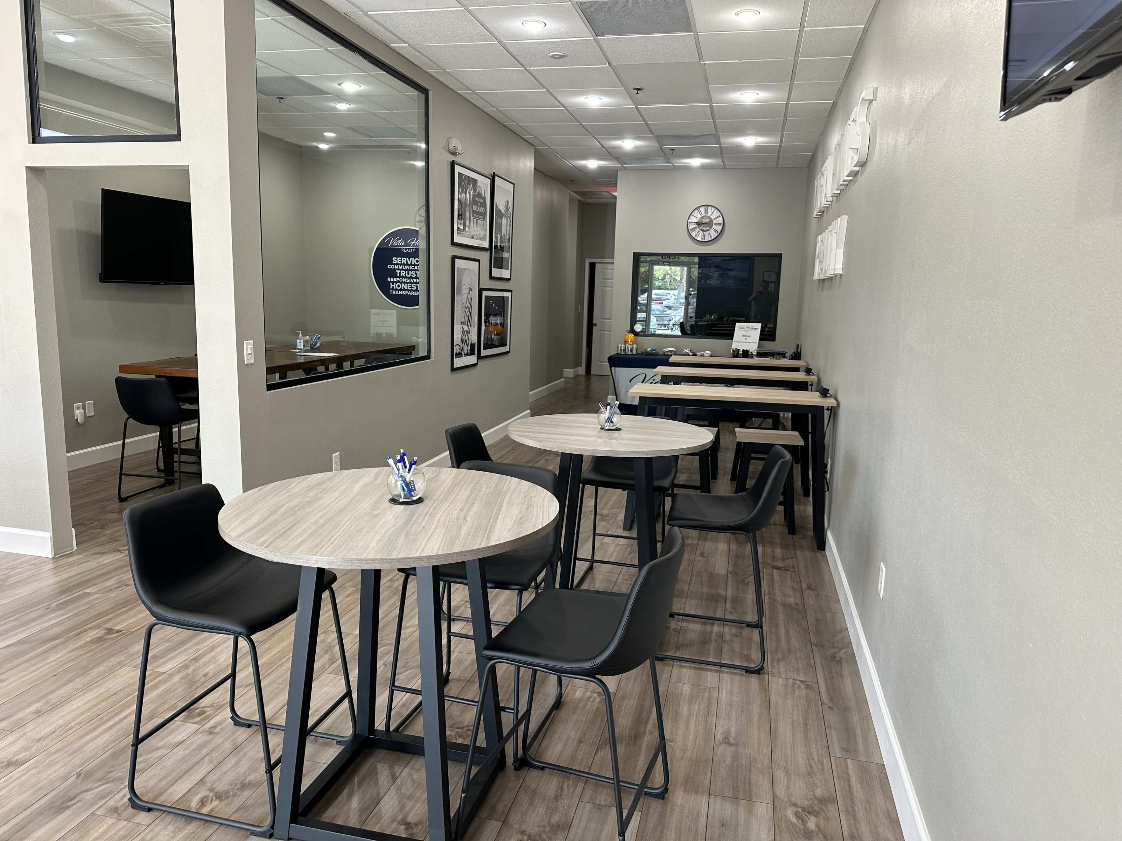 The image shows a modern, minimalistic interior of a cafe or office space featuring small round tables and black chairs, with a large window and various wall decorations.