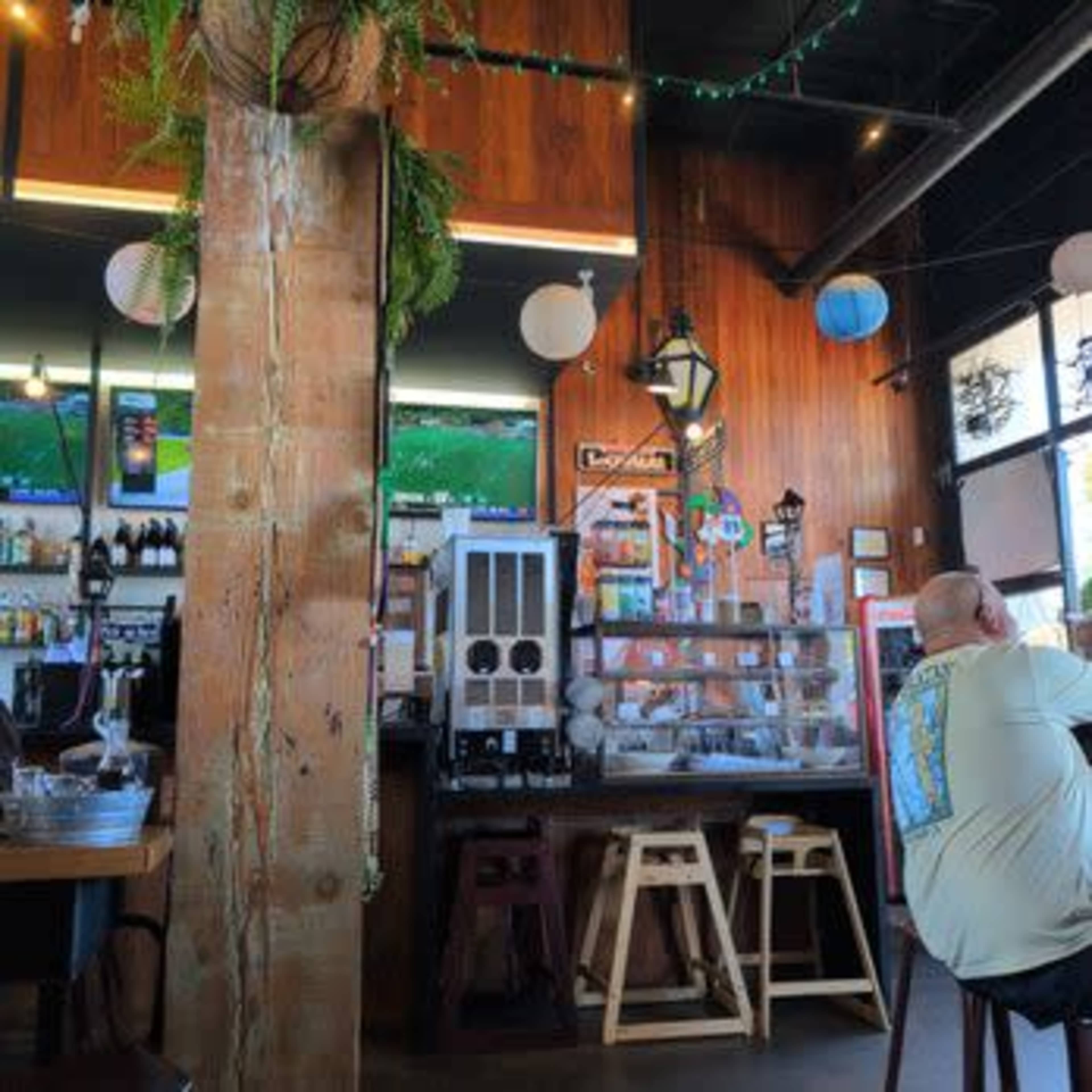 The image shows the interior of a casual bar or café with wooden walls, a counter displaying beverages, and a few patrons seated at tables.