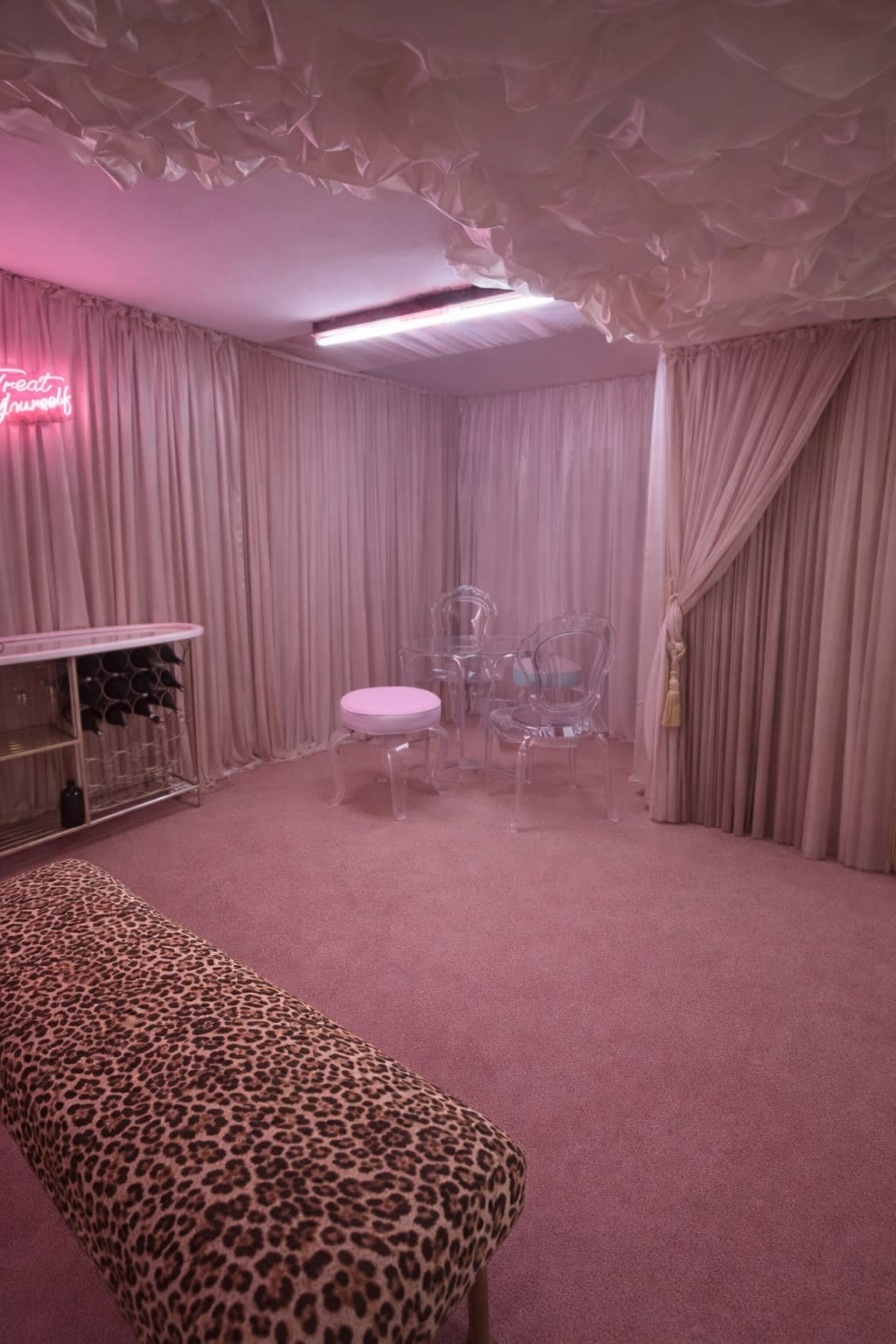 The room features pink walls and curtains, a leopard-print bench, a small circular pink table, and transparent chairs under soft lighting with a neon sign.