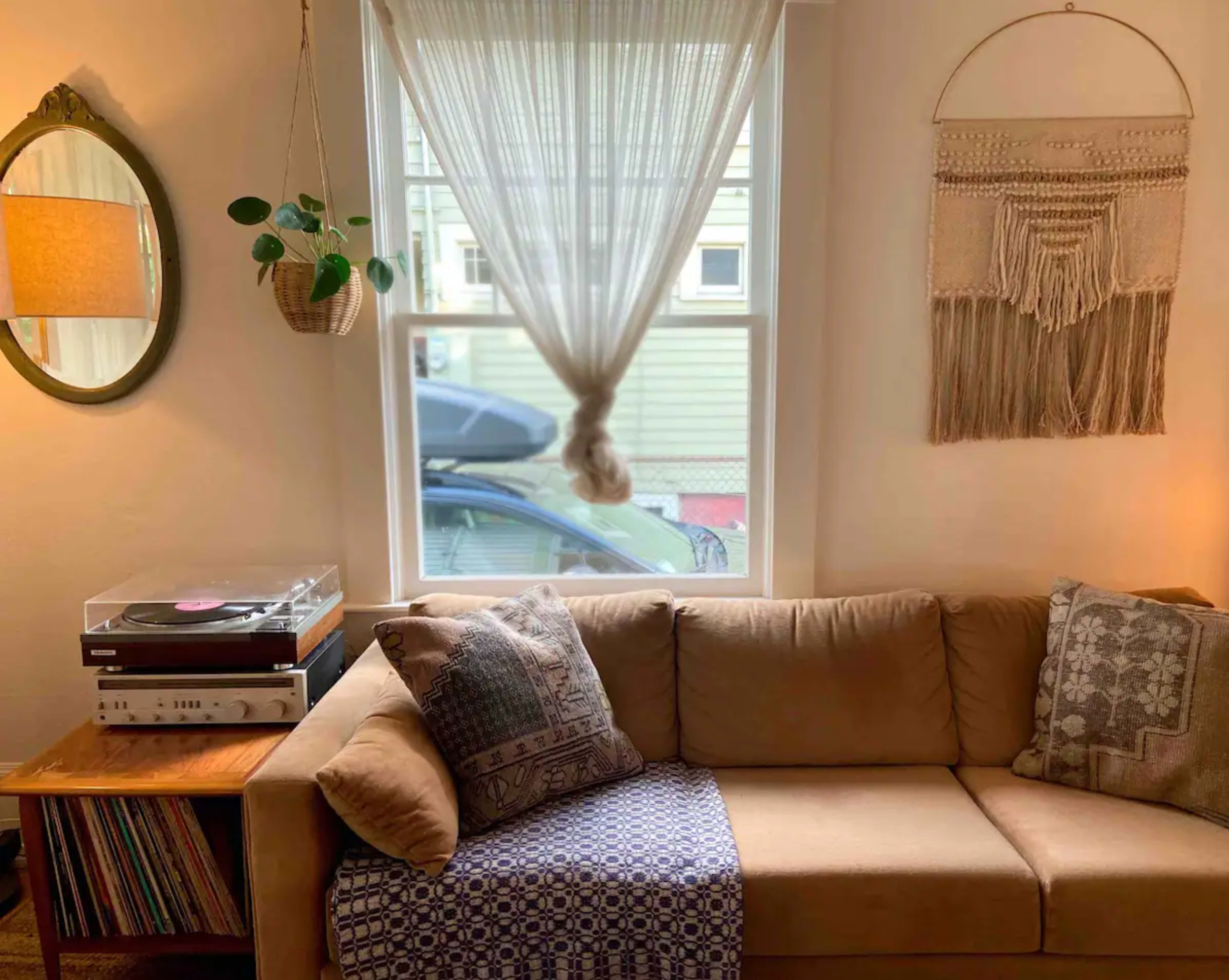 A cozy living room corner with a beige sofa, a decorative wall hanging, a window covered by sheer curtains, and a small record player next to a collection of vinyl records.