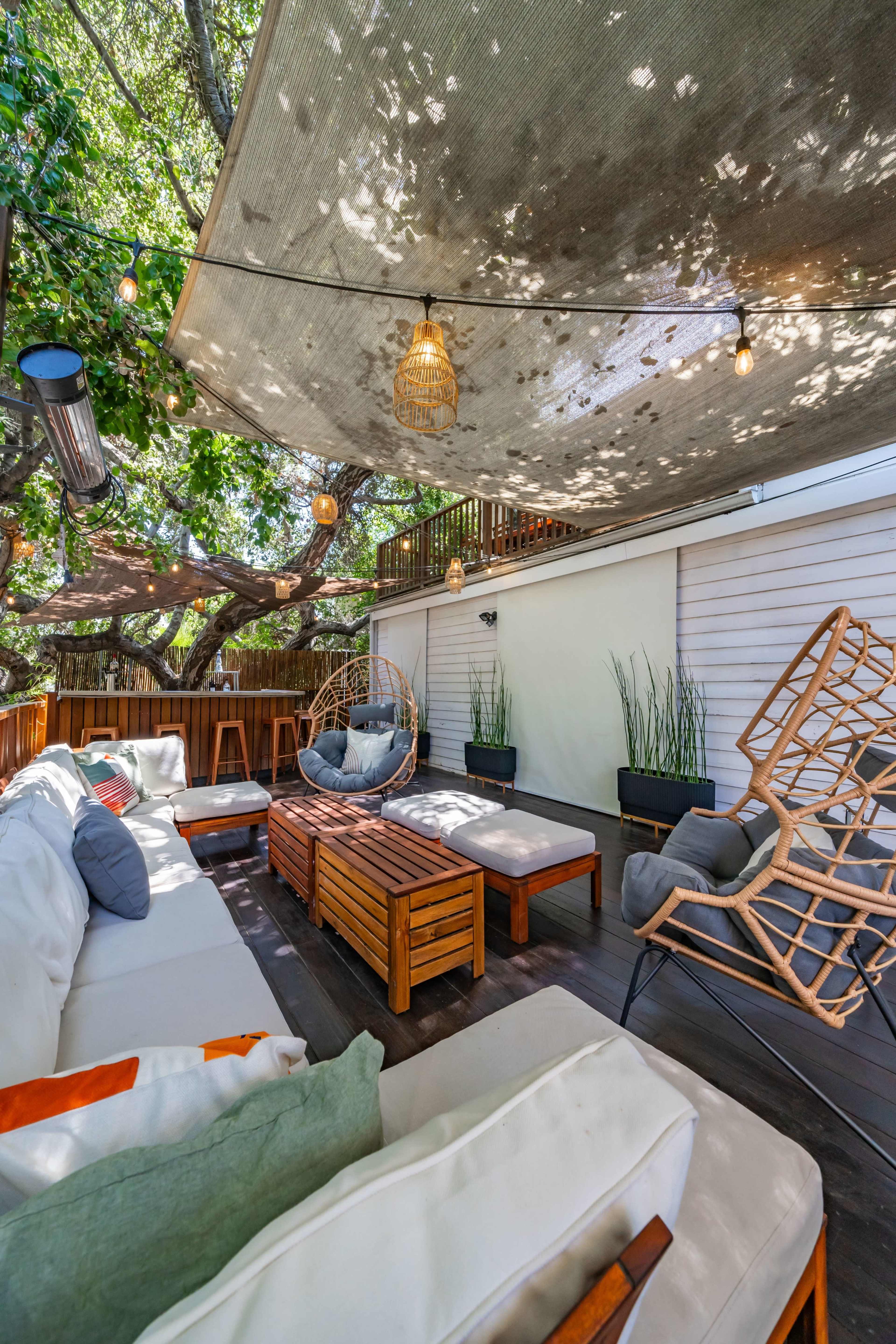 The image shows a cozy outdoor seating area with a shaded canopy, wooden furniture, and decorative plants.