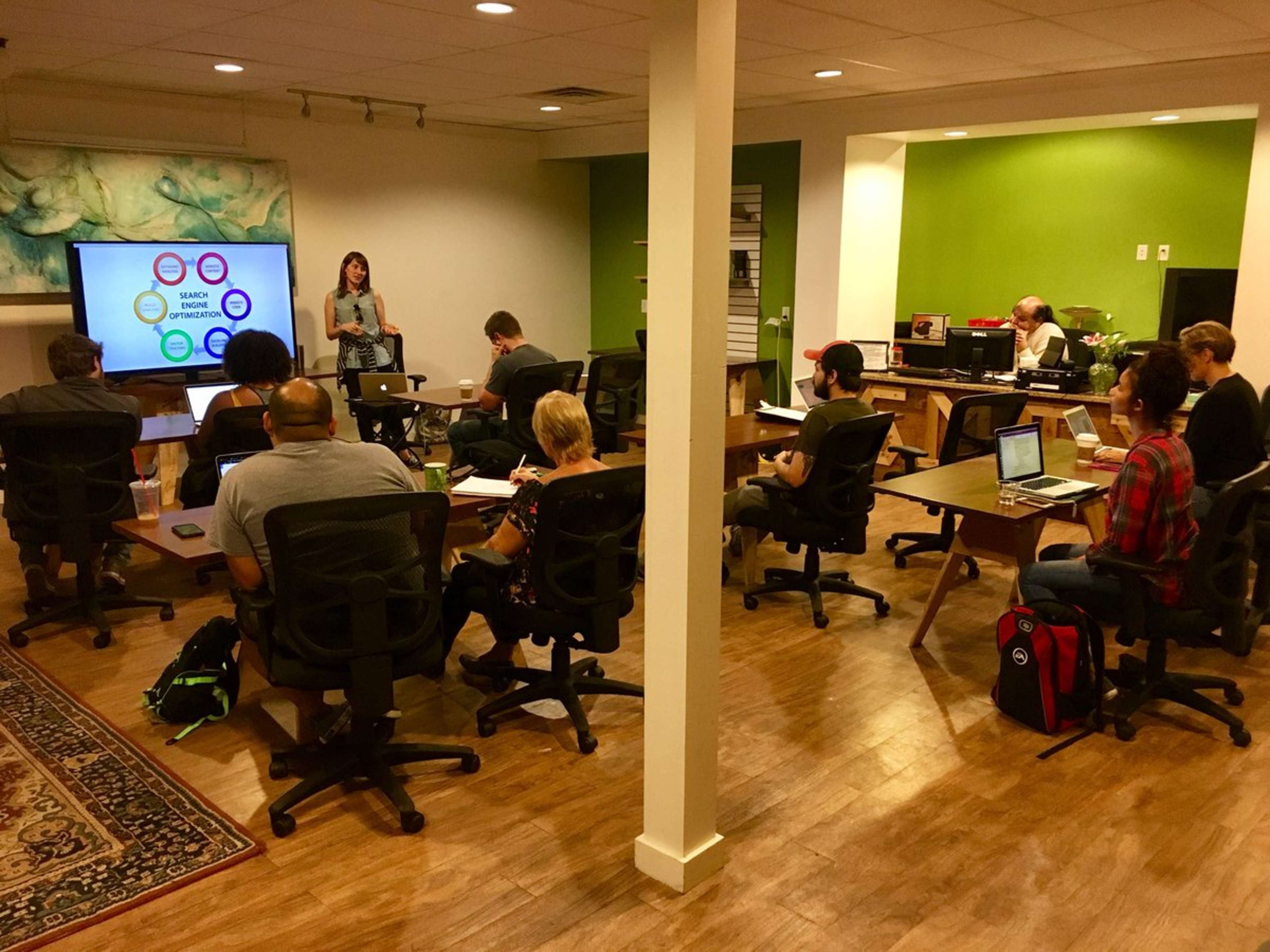 A woman presents information on a digital screen to an audience seated at tables in a well-lit classroom with green walls.