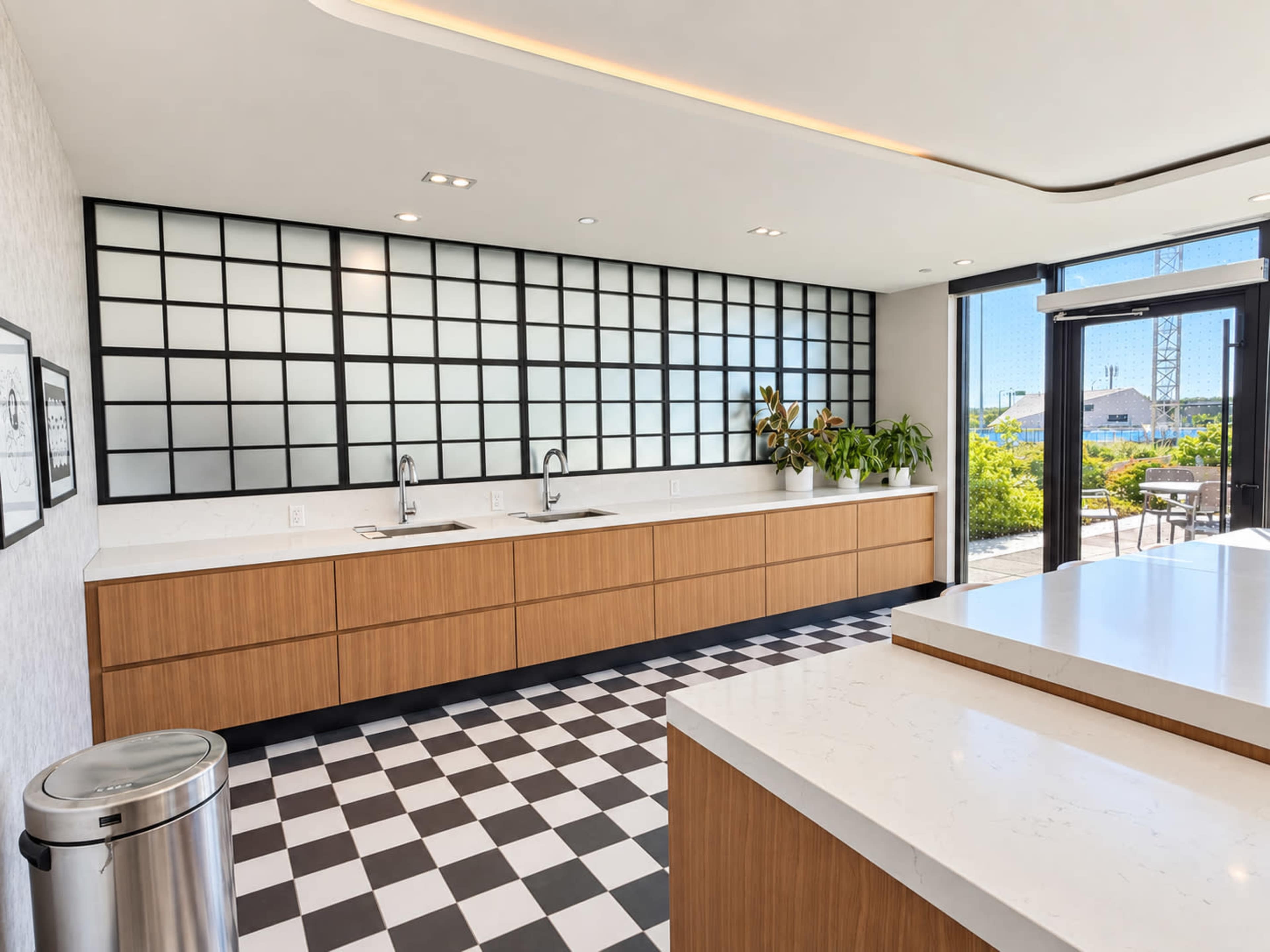 The image shows a modern kitchen with a black and white checkered floor, wooden cabinetry, and a large window overlooking greenery.