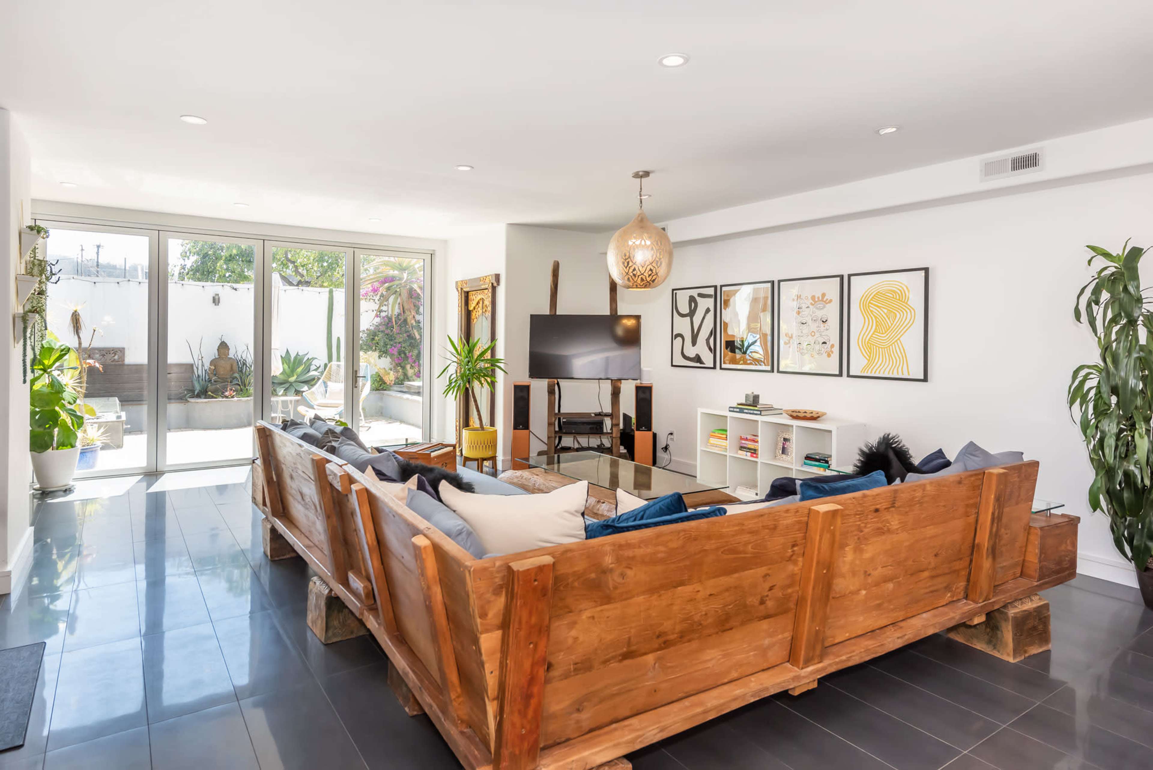 A modern living room featuring a large wooden sectional sofa, a flat-screen TV, and framed artwork on the walls, with large windows providing natural light and a view of an outdoor area.