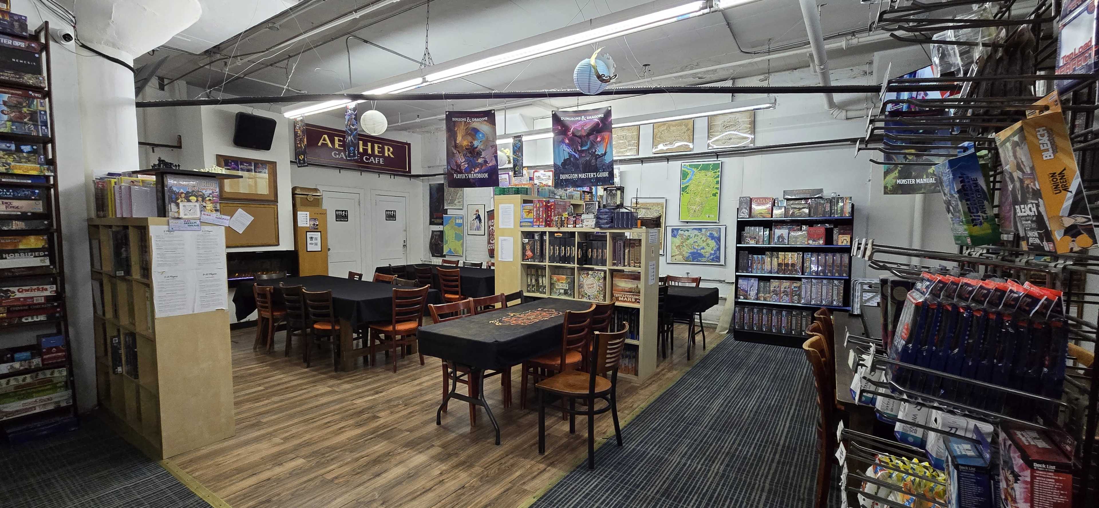 The image shows a spacious game cafe with tables set for board games, shelves filled with game boxes, and posters on the walls.