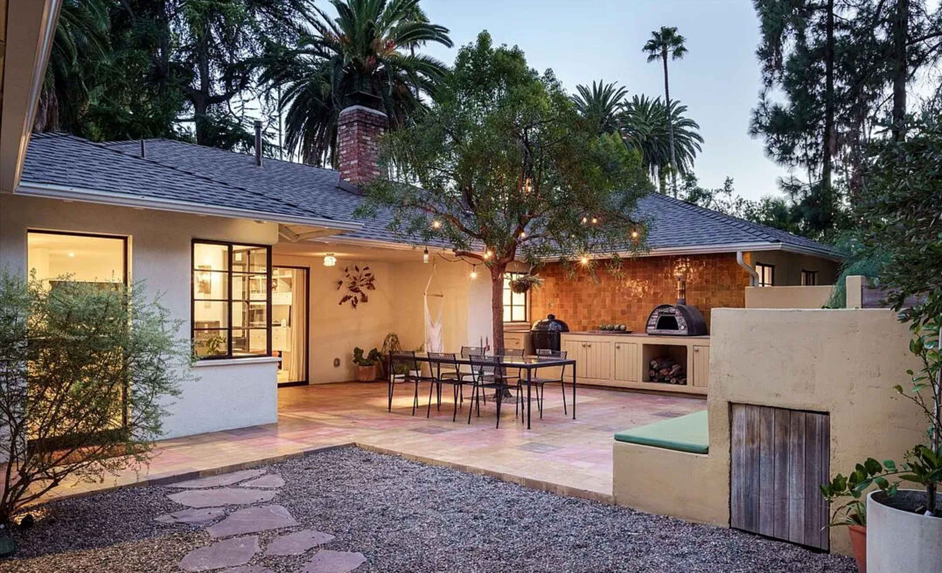 A patio area features a dining table set under a tree with string lights, alongside an outdoor kitchen and a pizza oven, surrounded by landscaping and palm trees.