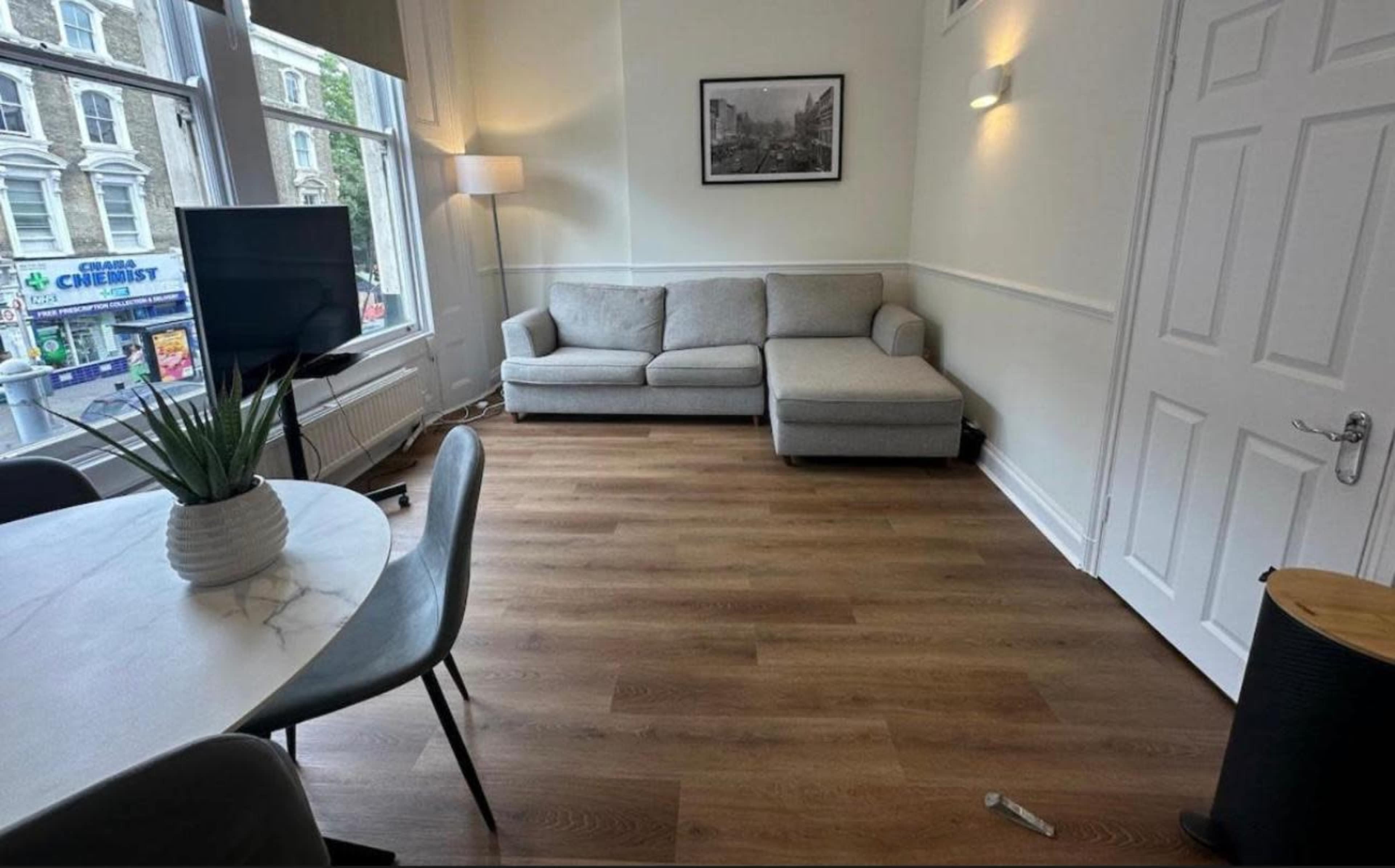 The image shows a living area with a gray sectional sofa, a round dining table with chairs, a television, and wooden flooring.