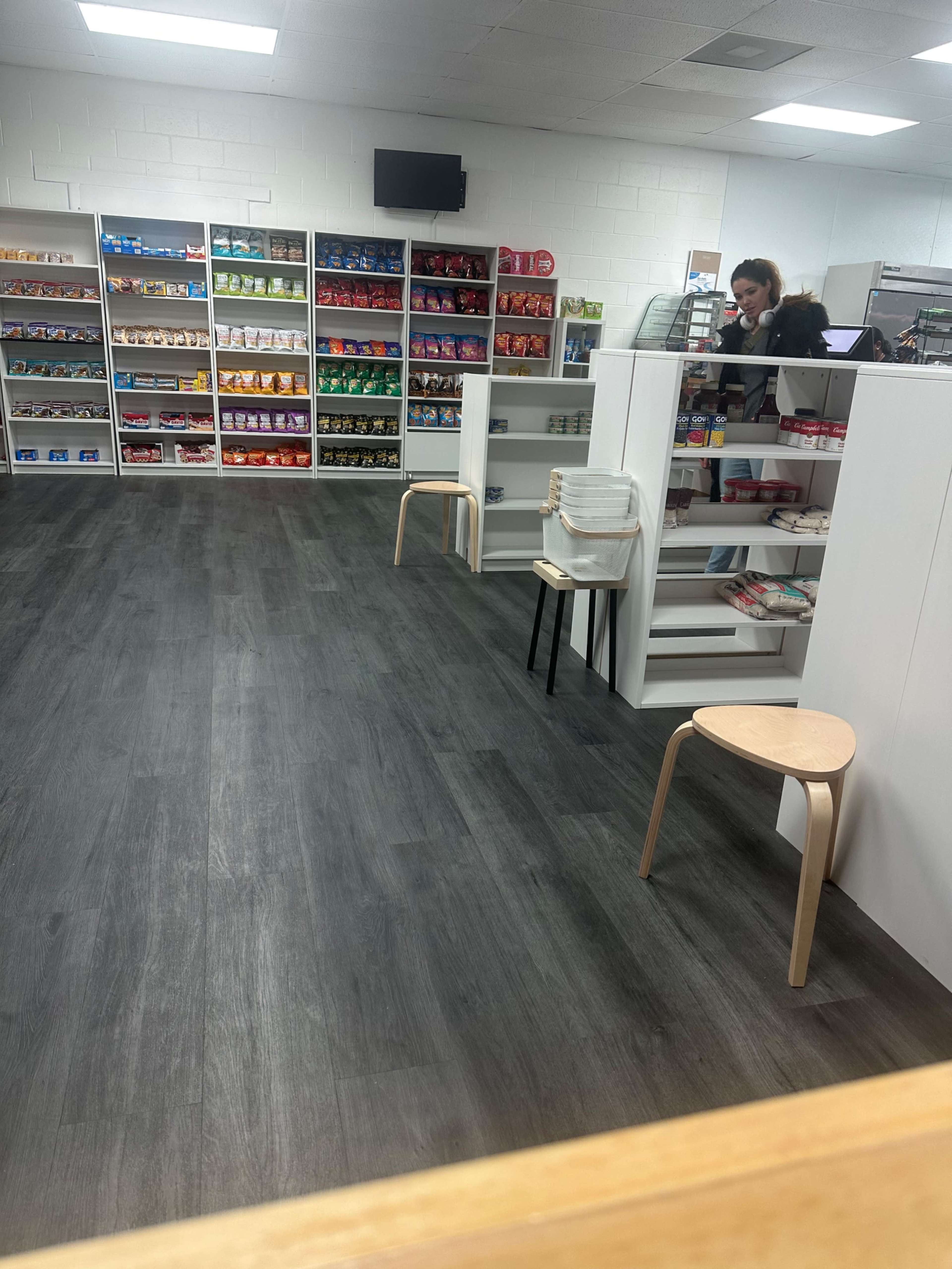 A well-organized retail space features shelves filled with colorful products, with a woman standing behind a counter and empty chairs nearby.