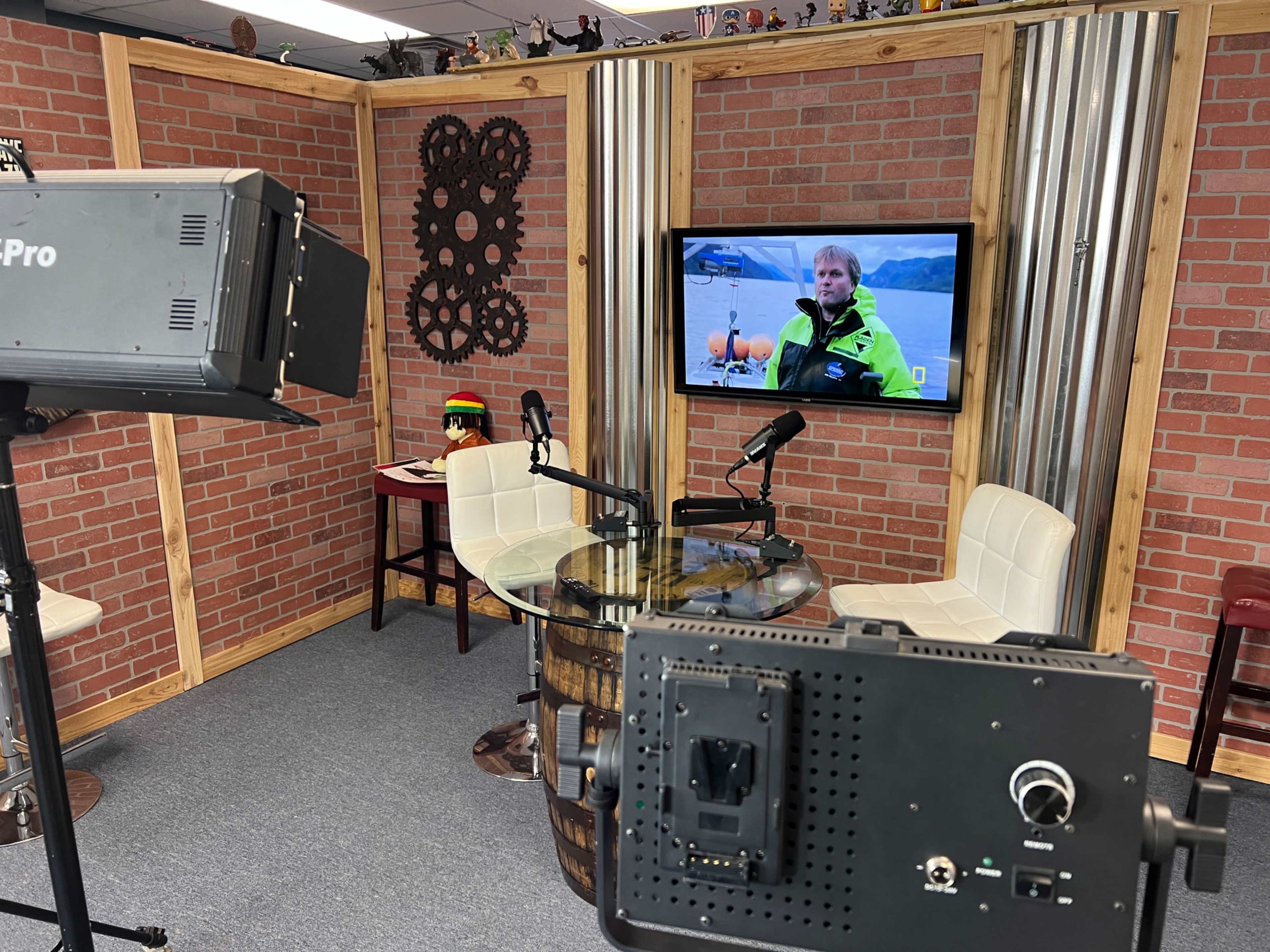 The image shows a small studio setup with a glass table, two white chairs, and a television displaying a man in a yellow jacket, framed by a brick wall and decorative elements.