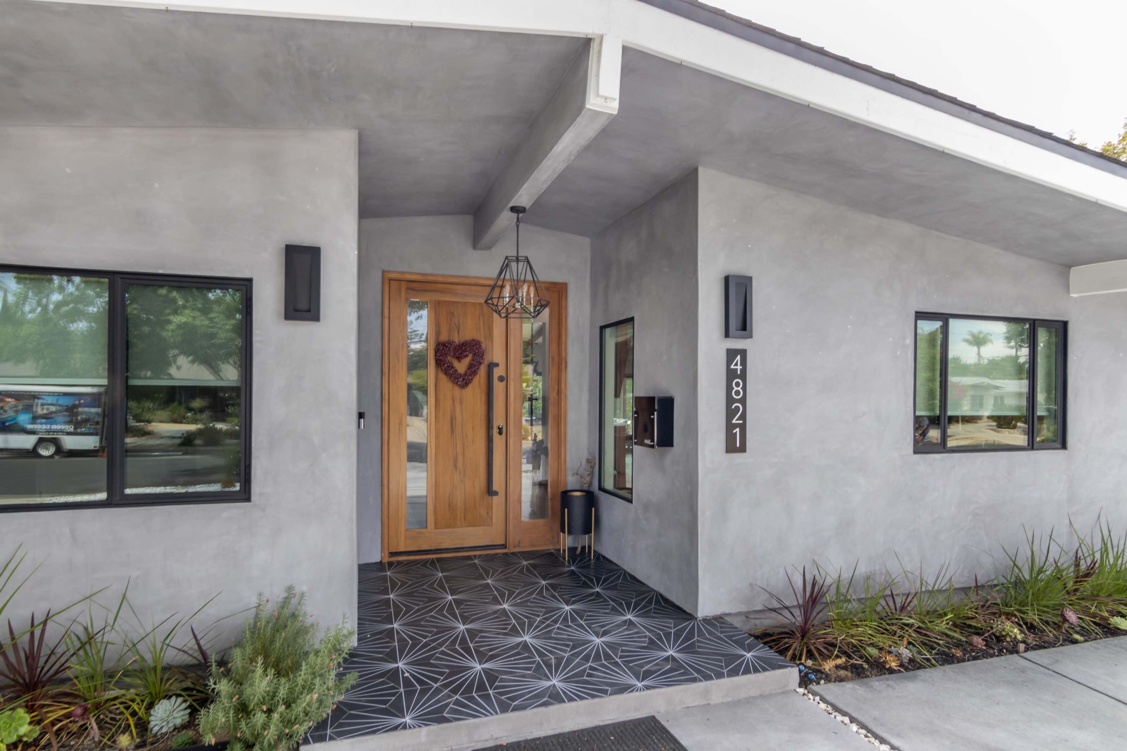 The entrance of a modern home featuring a wooden door, gray concrete walls, and a decorative tile pathway.