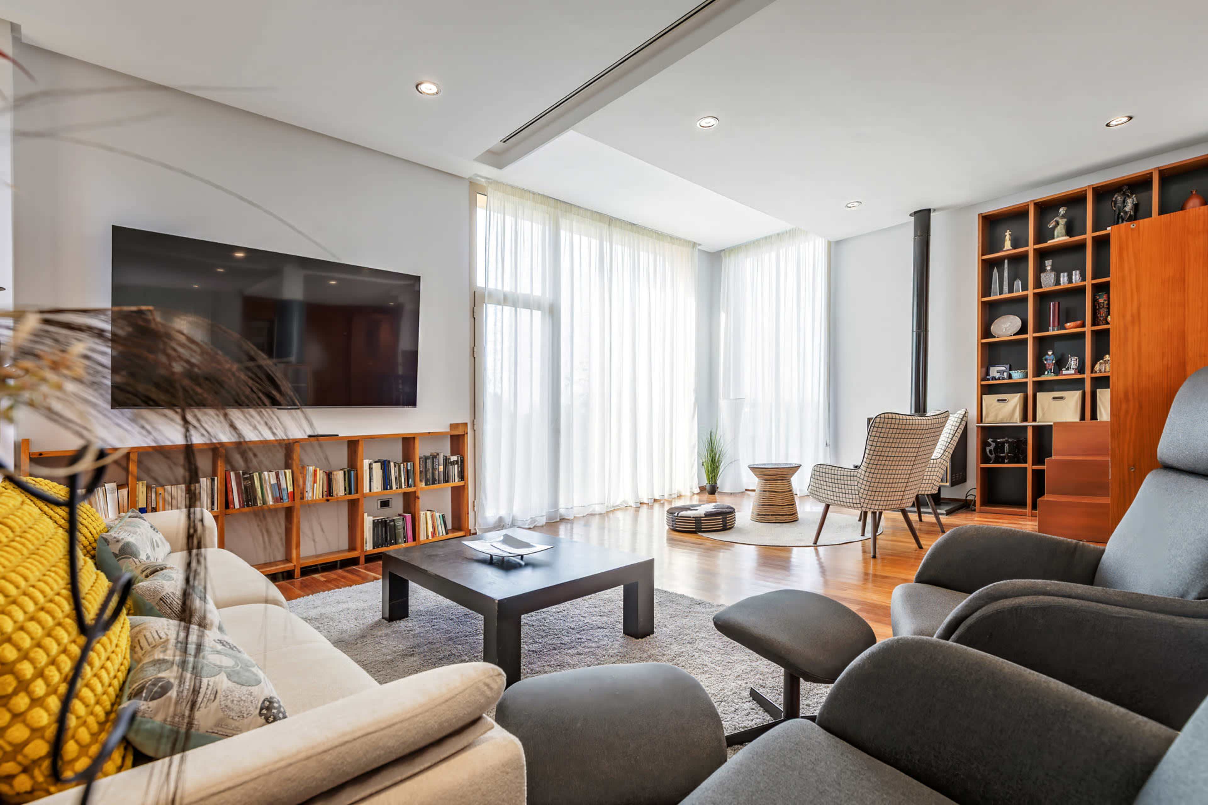 The image shows a modern living room with a large TV, a comfortable seating area, a bookshelf, and large windows with sheer curtains.