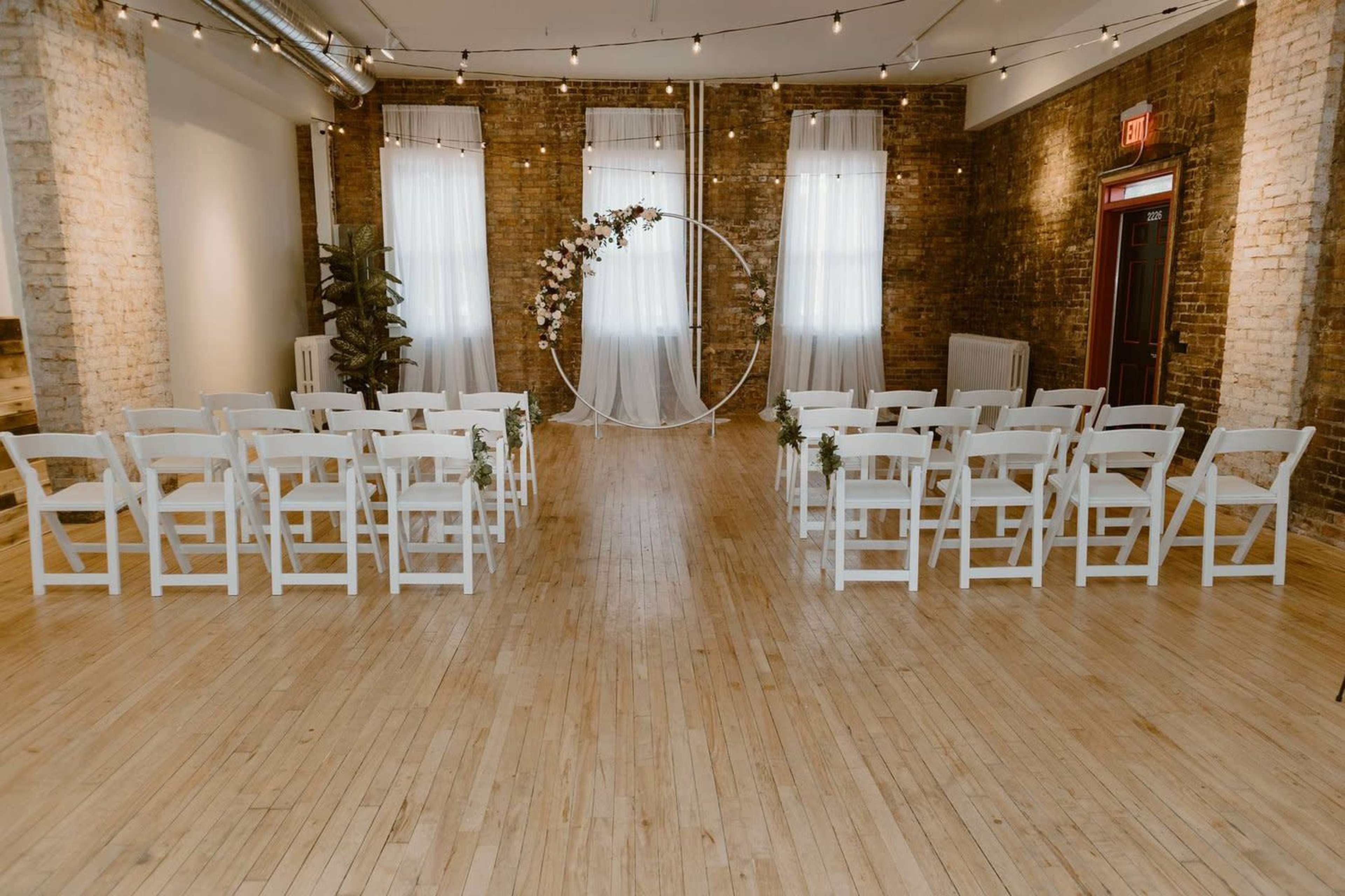 A spacious venue features neatly arranged white chairs facing a circular floral arch, with large windows and exposed brick walls in the background.