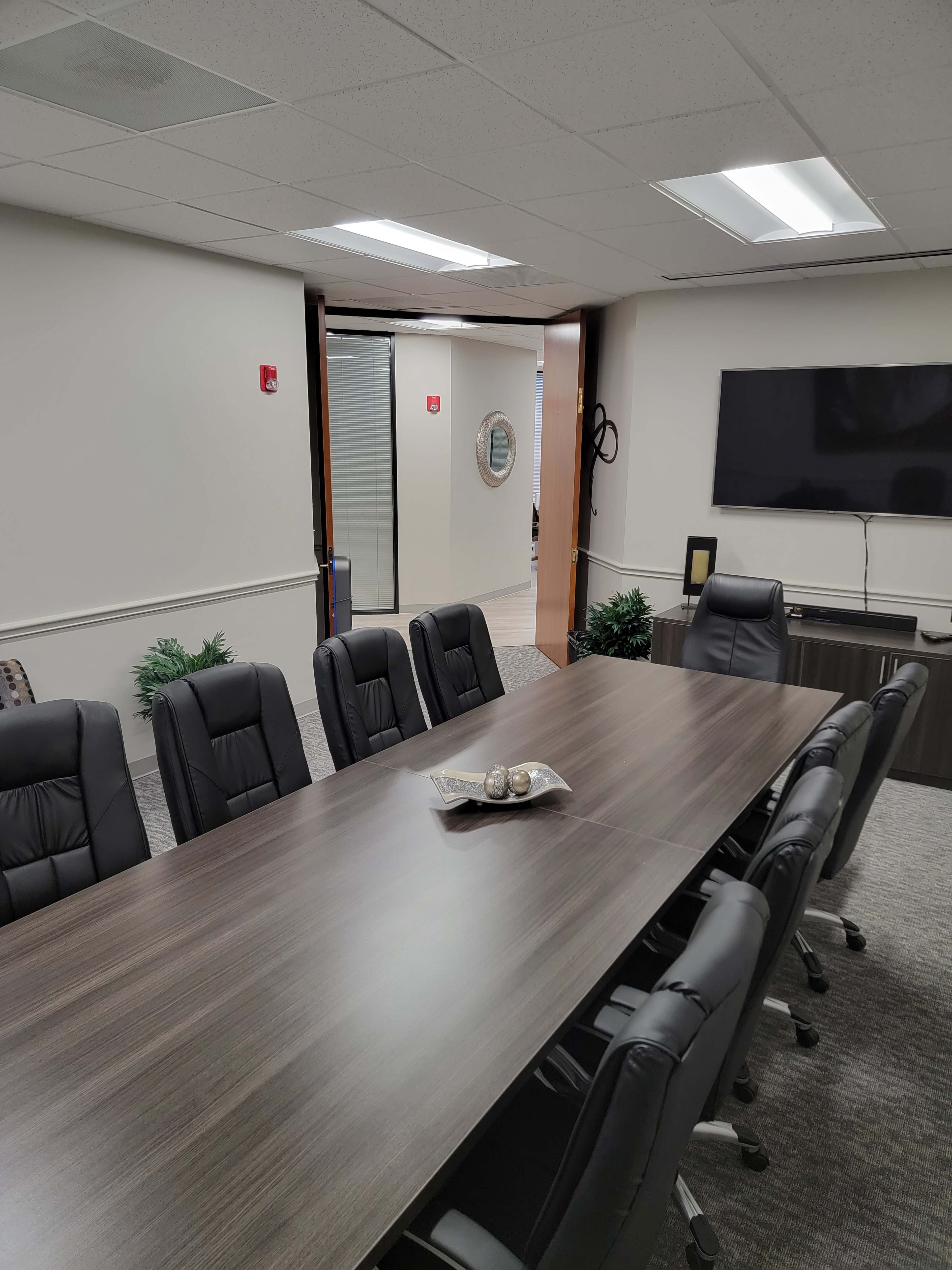 The image shows a modern conference room with a long wooden table surrounded by black chairs, leading to a glass door that opens to a hallway.