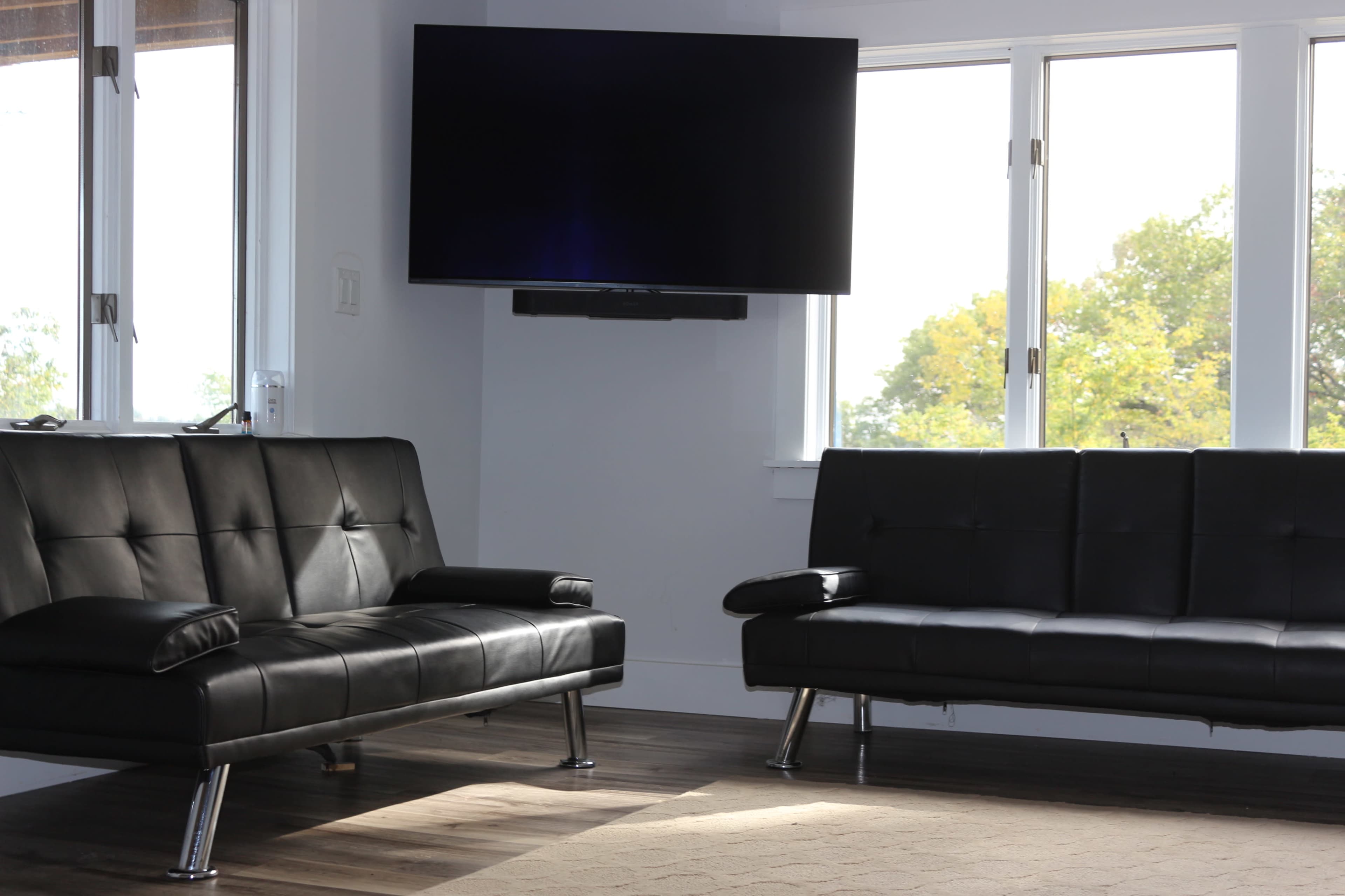 A living room with two black sofas facing each other, a wall-mounted television, and large windows that allow natural light to enter.