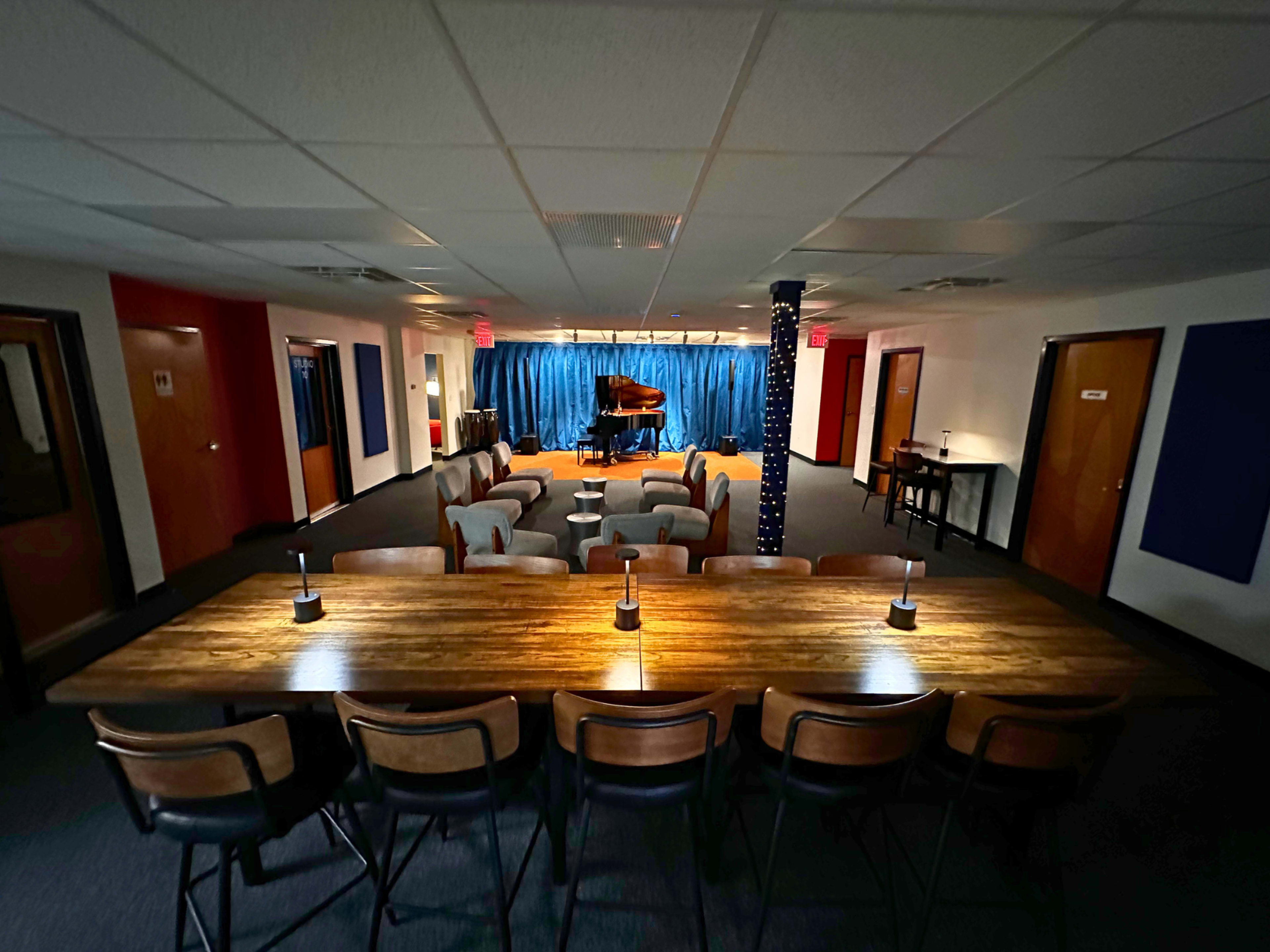 The image shows a spacious music lounge with a long wooden table and chairs in the foreground, facing a stage draped in blue fabric featuring a grand piano.