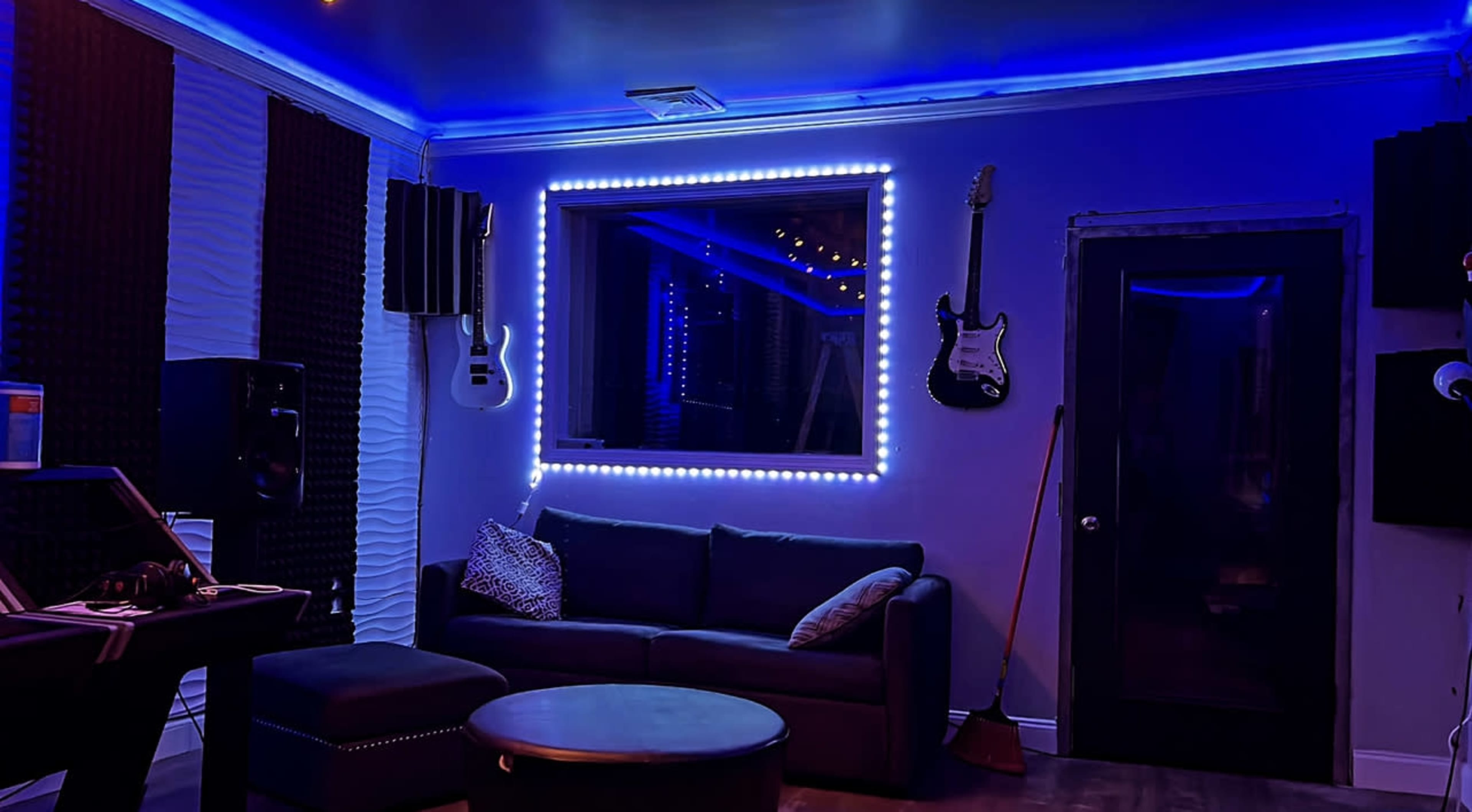 A music studio room with a dark sofa, a circular table, guitar hangers on the wall, soundproofing panels, and blue LED lighting around a window.