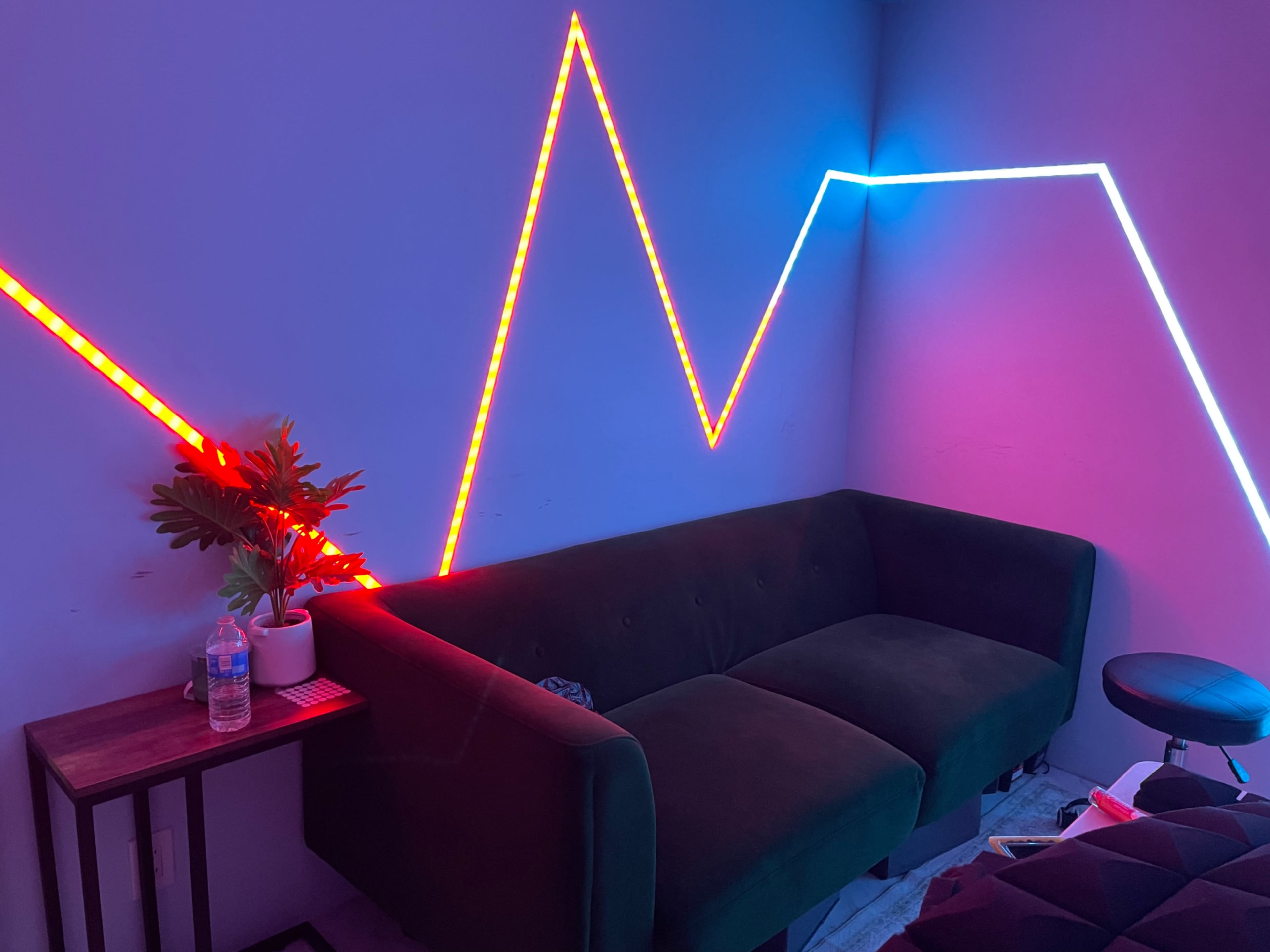 A dark room features a green sofa against a wall illuminated by red and blue LED light strips arranged in a zigzag pattern, with a small table and a potted plant nearby.