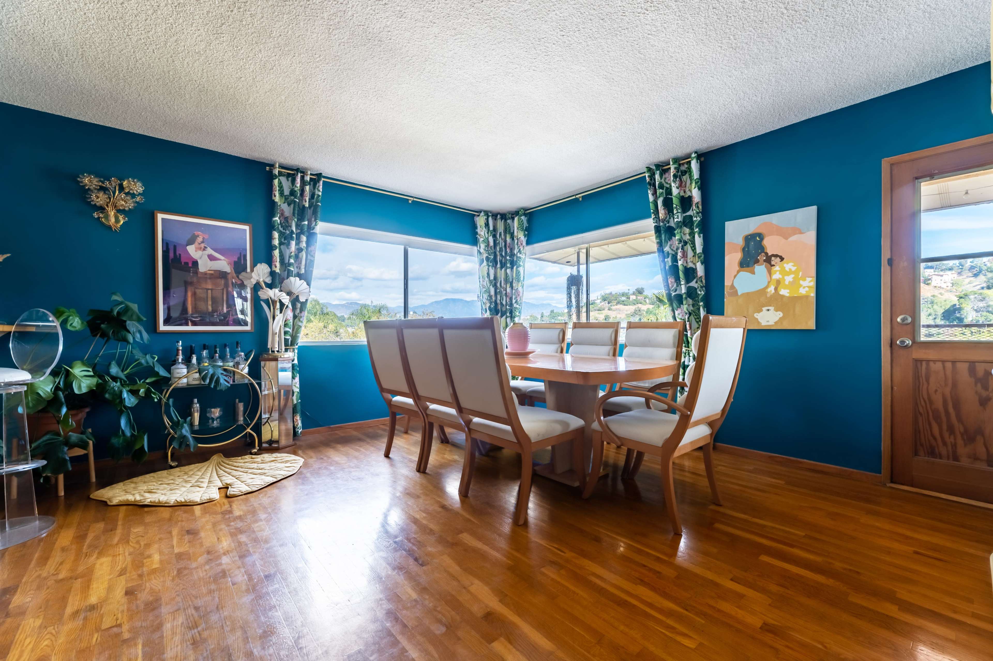 The image shows a dining room with a wooden table and chairs, large windows providing natural light, and teal walls decorated with artwork and plants.