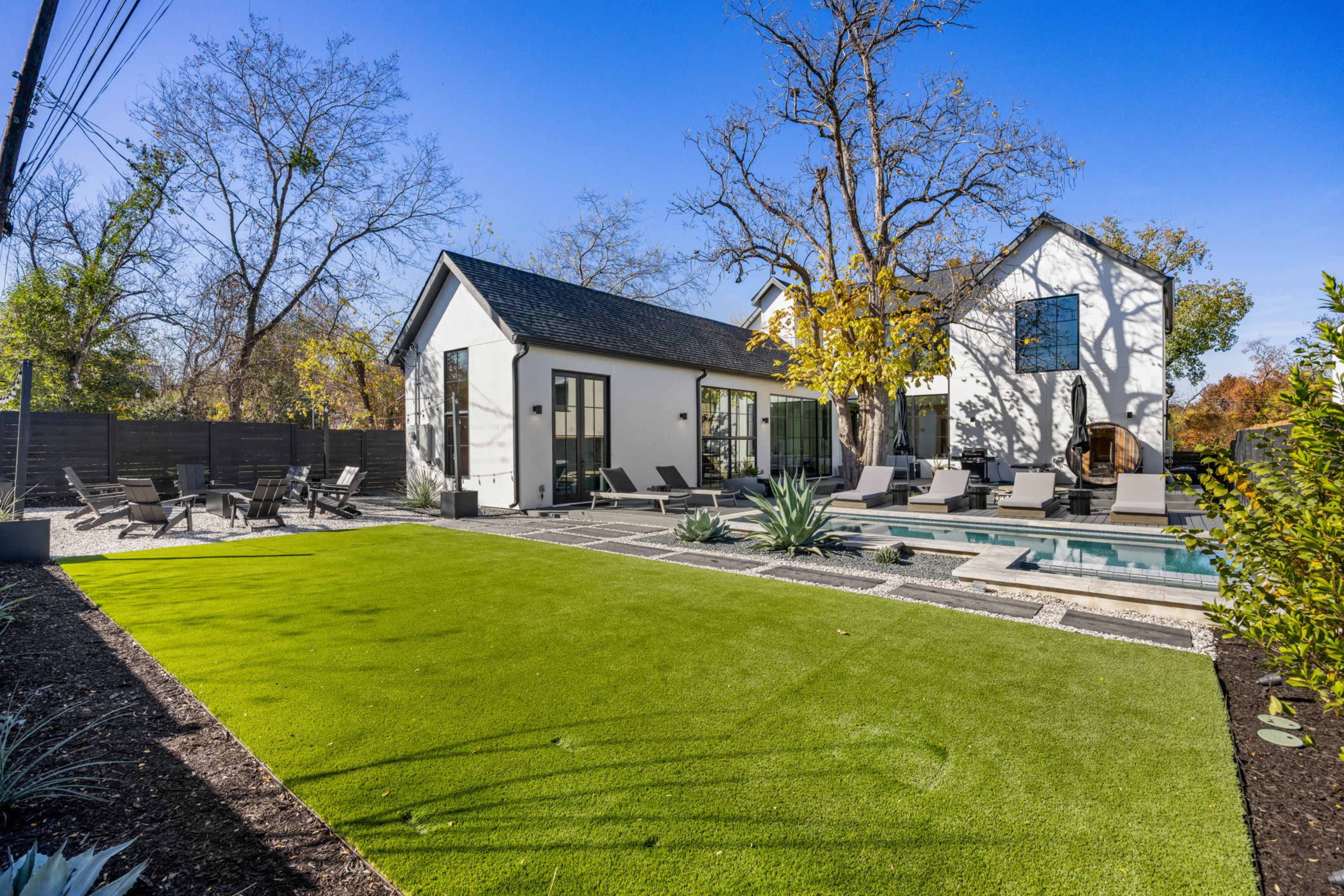 The image shows a modern residential backyard featuring a swimming pool, lounge chairs, and a well-maintained strip of artificial turf surrounded by trees and landscaping.