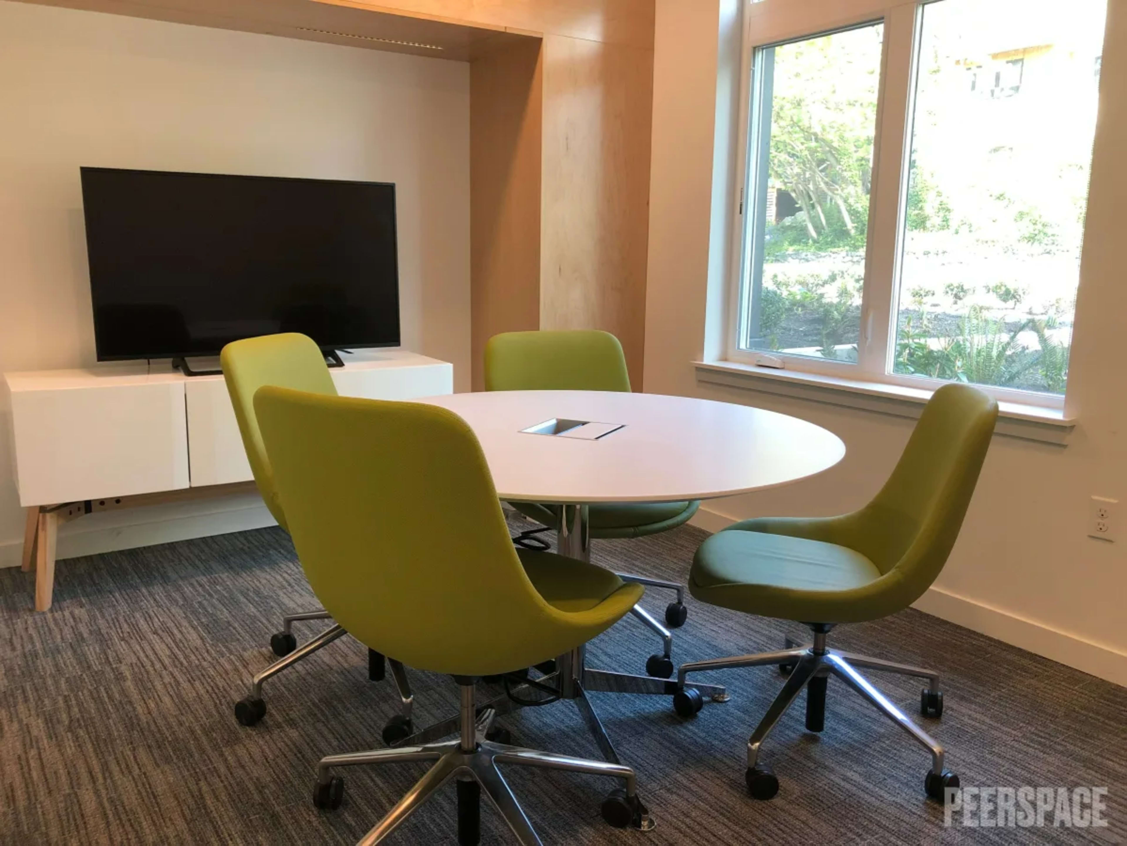 The image shows a modern meeting room with a round table surrounded by four green chairs and a television mounted on the wall.