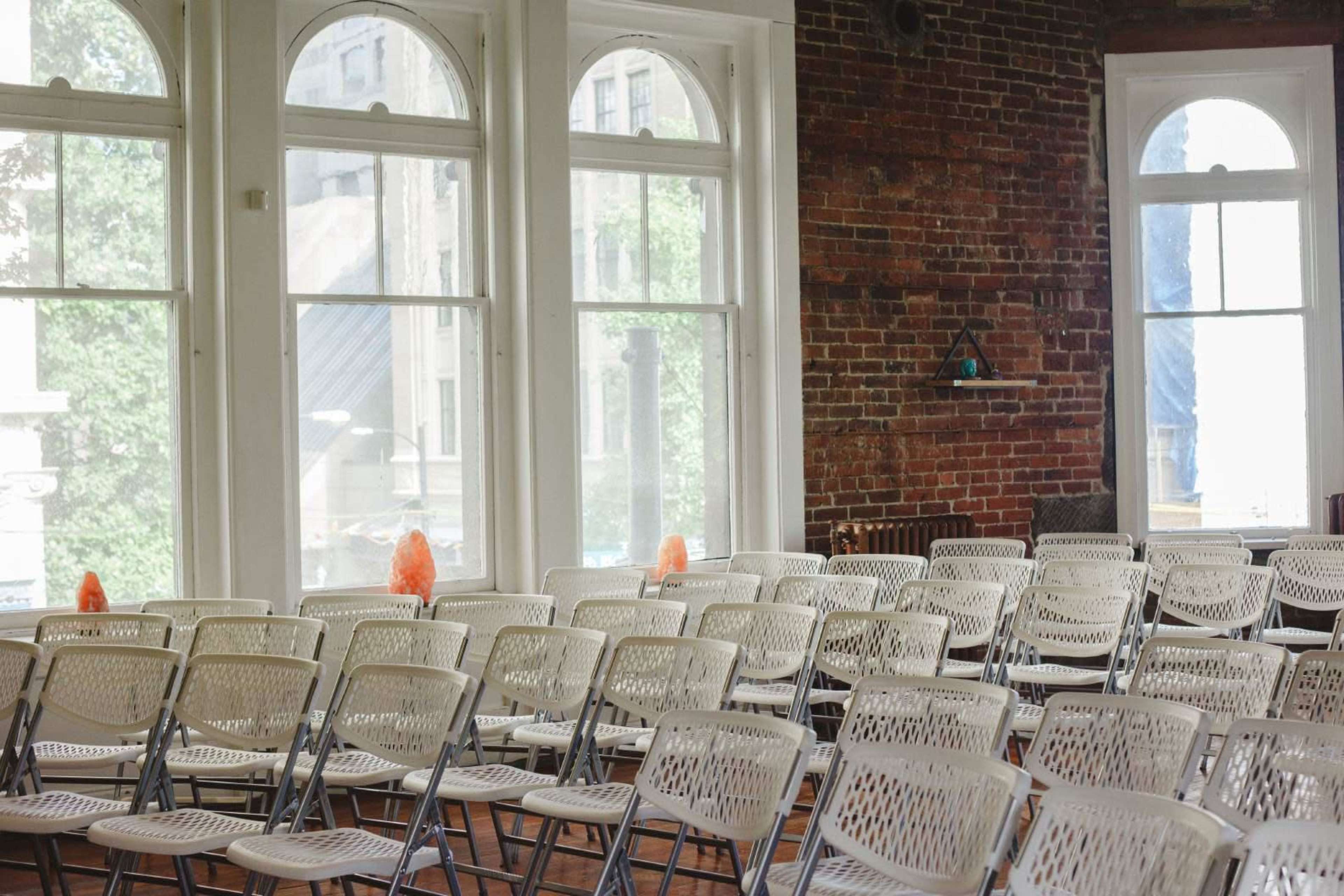 A row of white folding chairs arranged in a room with large windows and exposed brick walls.