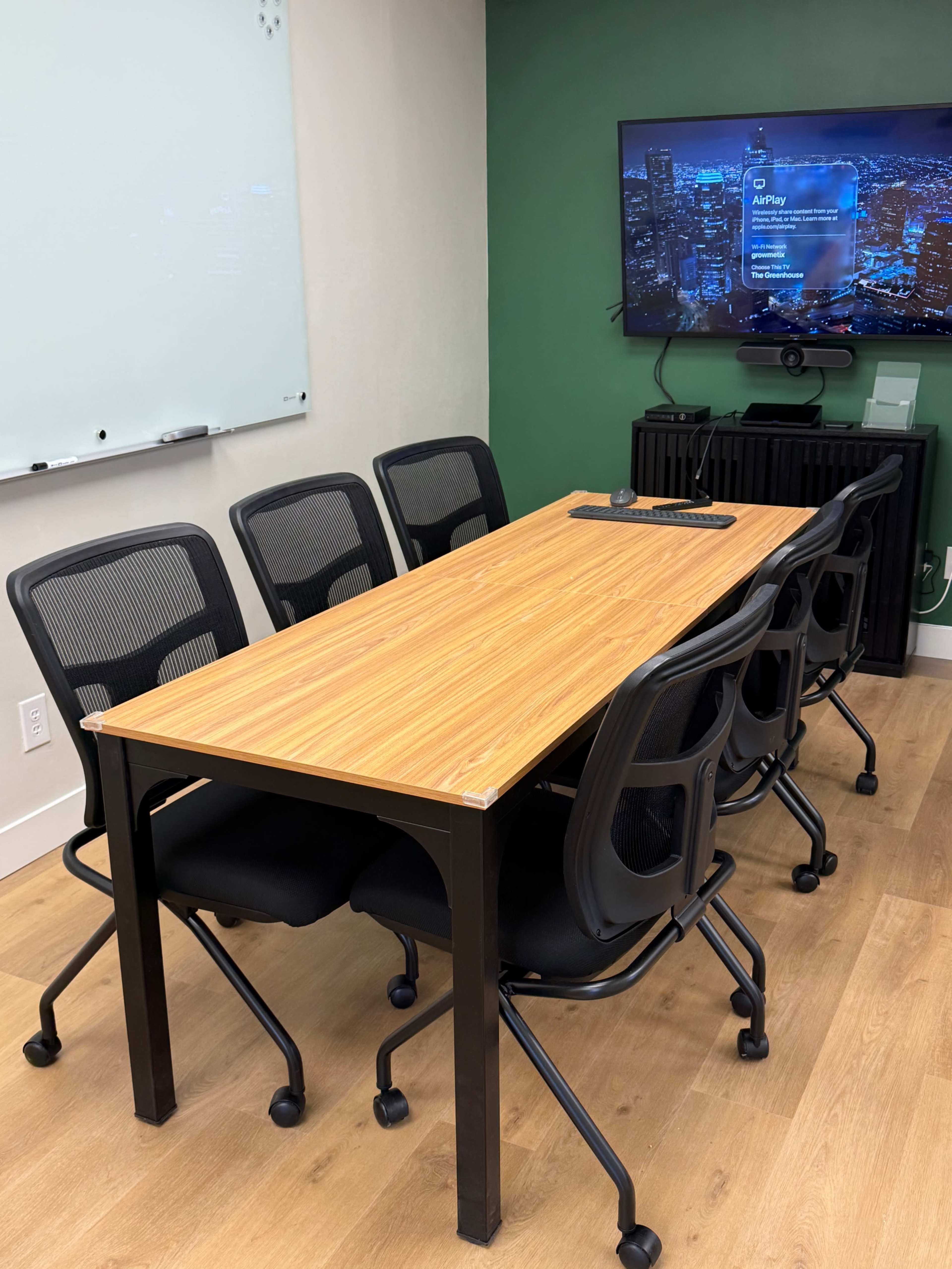A conference room features a wooden table surrounded by six black rolling chairs, with a large screen displaying content on a green wall.