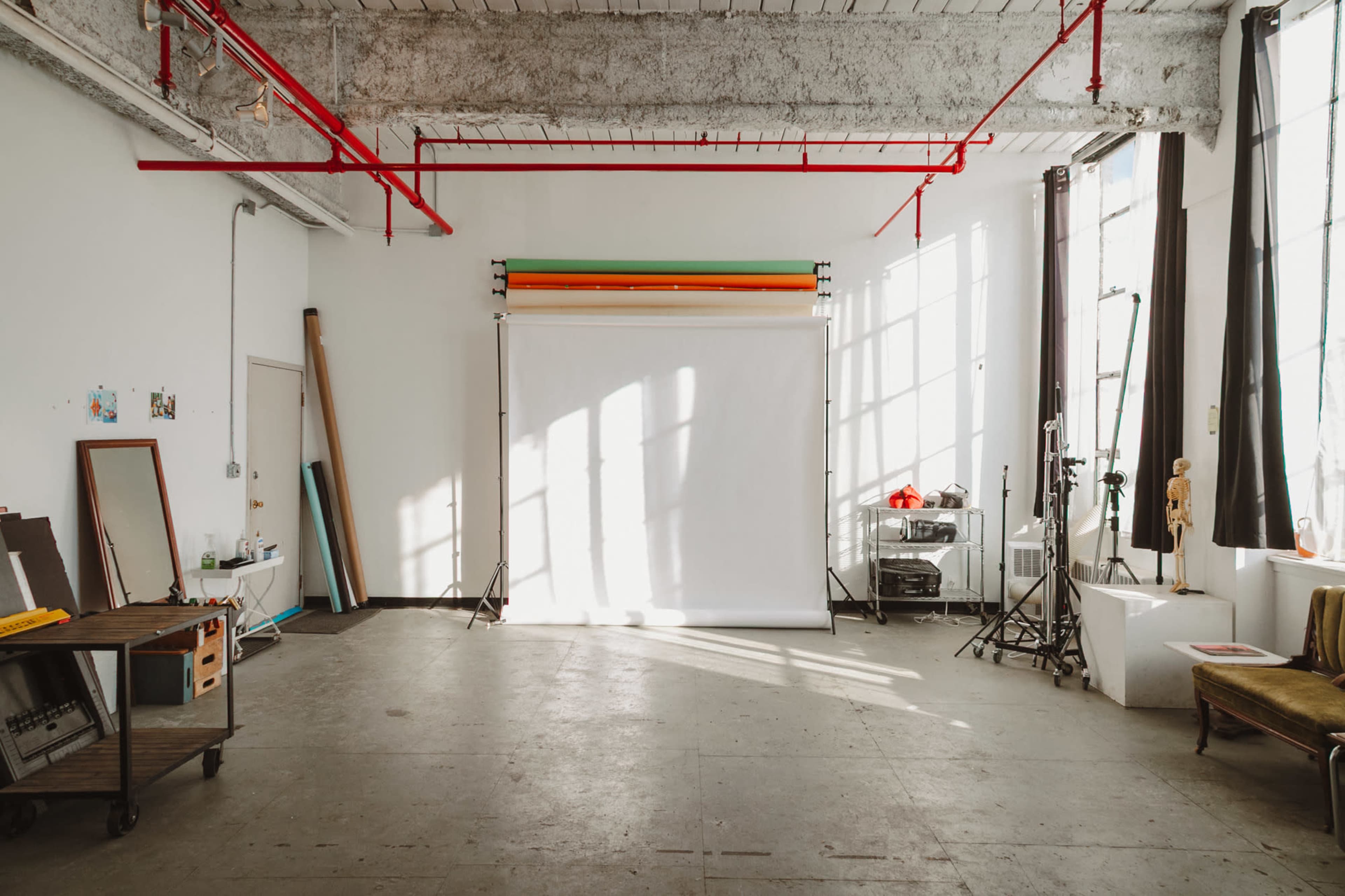 The image shows a spacious photography studio with large windows allowing natural light to illuminate a white backdrop, and various equipment and props arranged around the room.