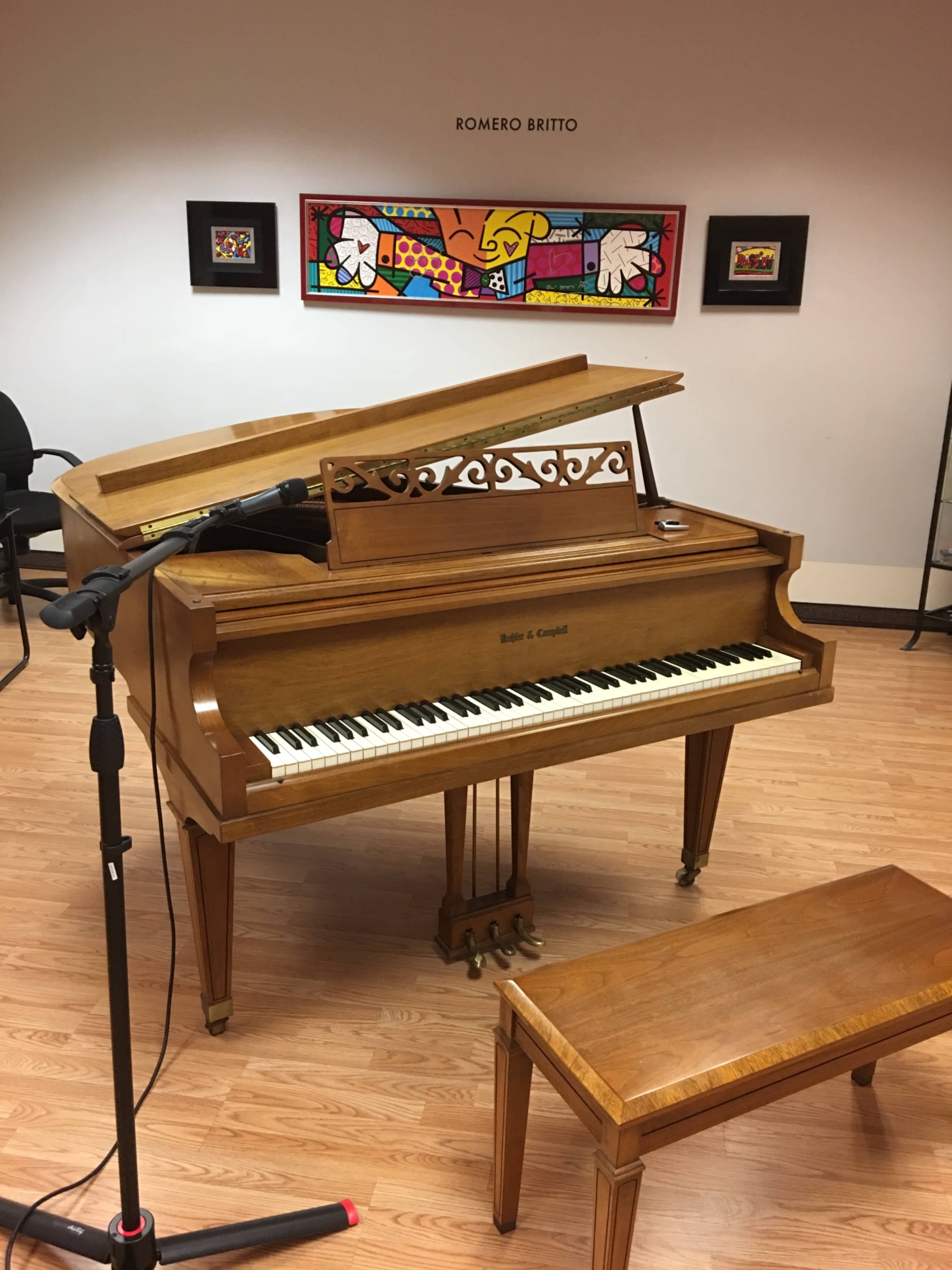 A wooden grand piano with an open lid stands in a room featuring colorful artwork on the walls.