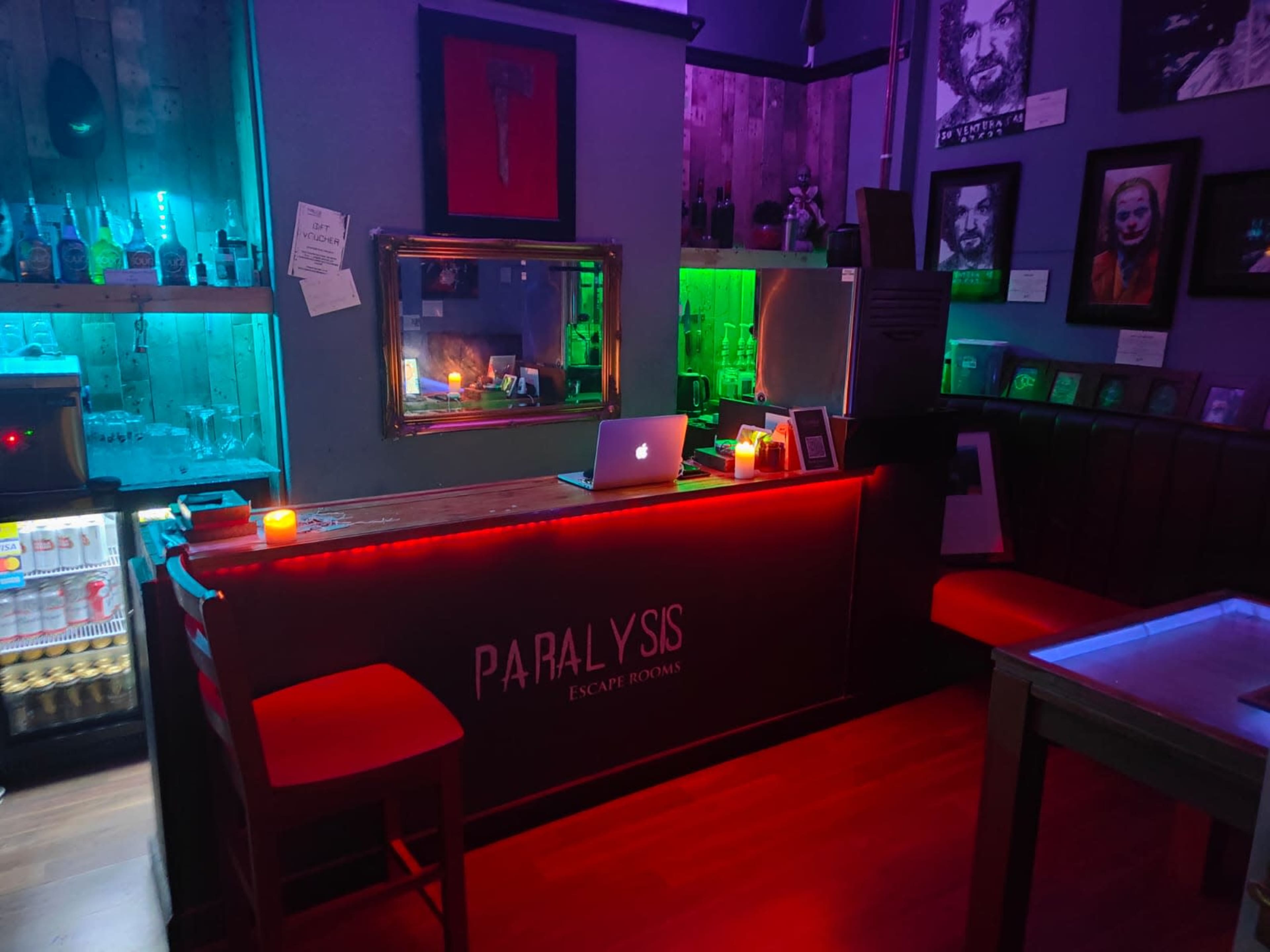 A dimly lit escape room bar with colorful neon lights, a sleek counter displaying the word "PARALYSIS," and various portraits on the walls.