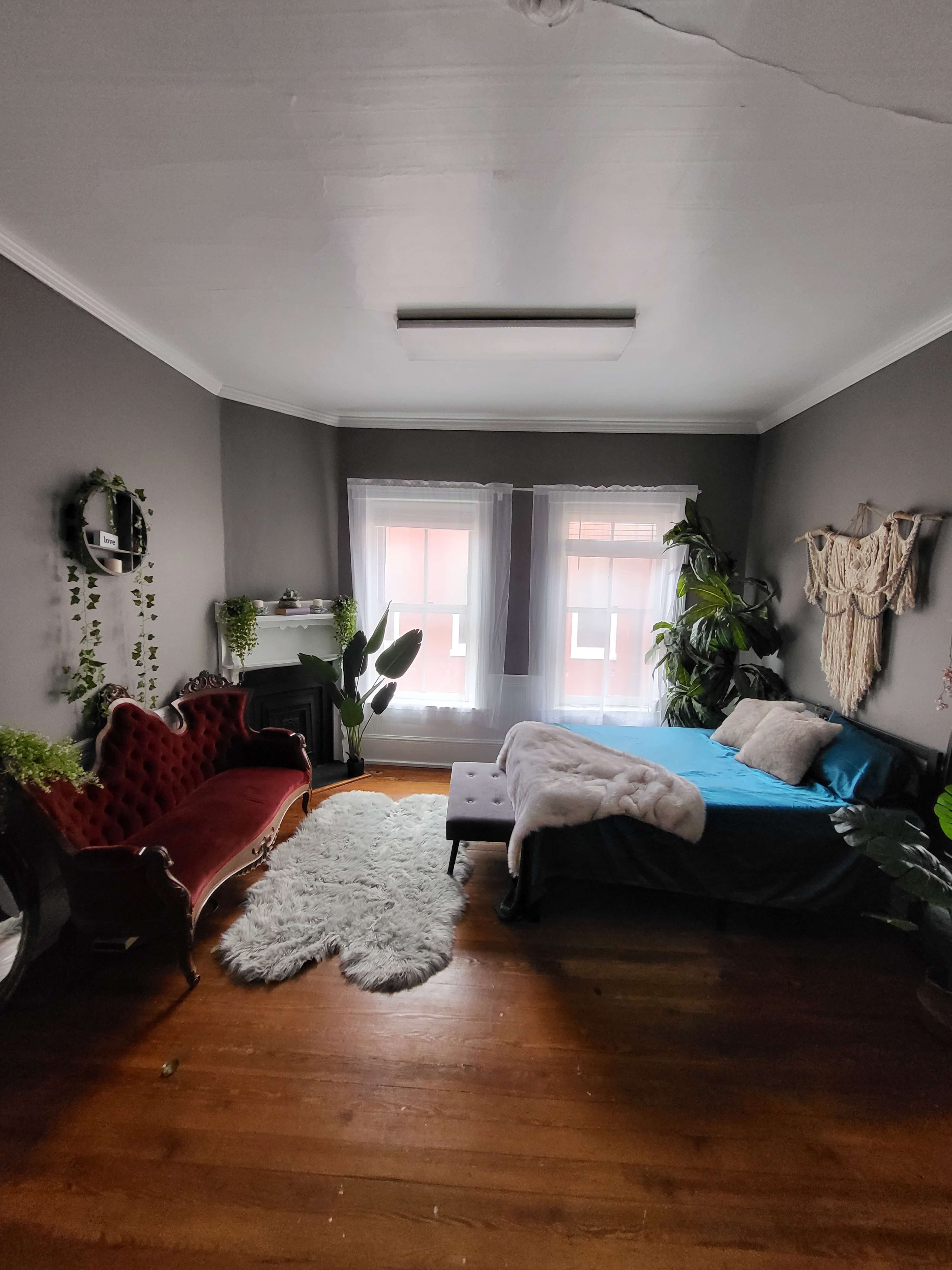 The image shows a cozy bedroom with a plush red velvet chair, a bed with blue bedding, houseplants, and large windows letting in natural light.