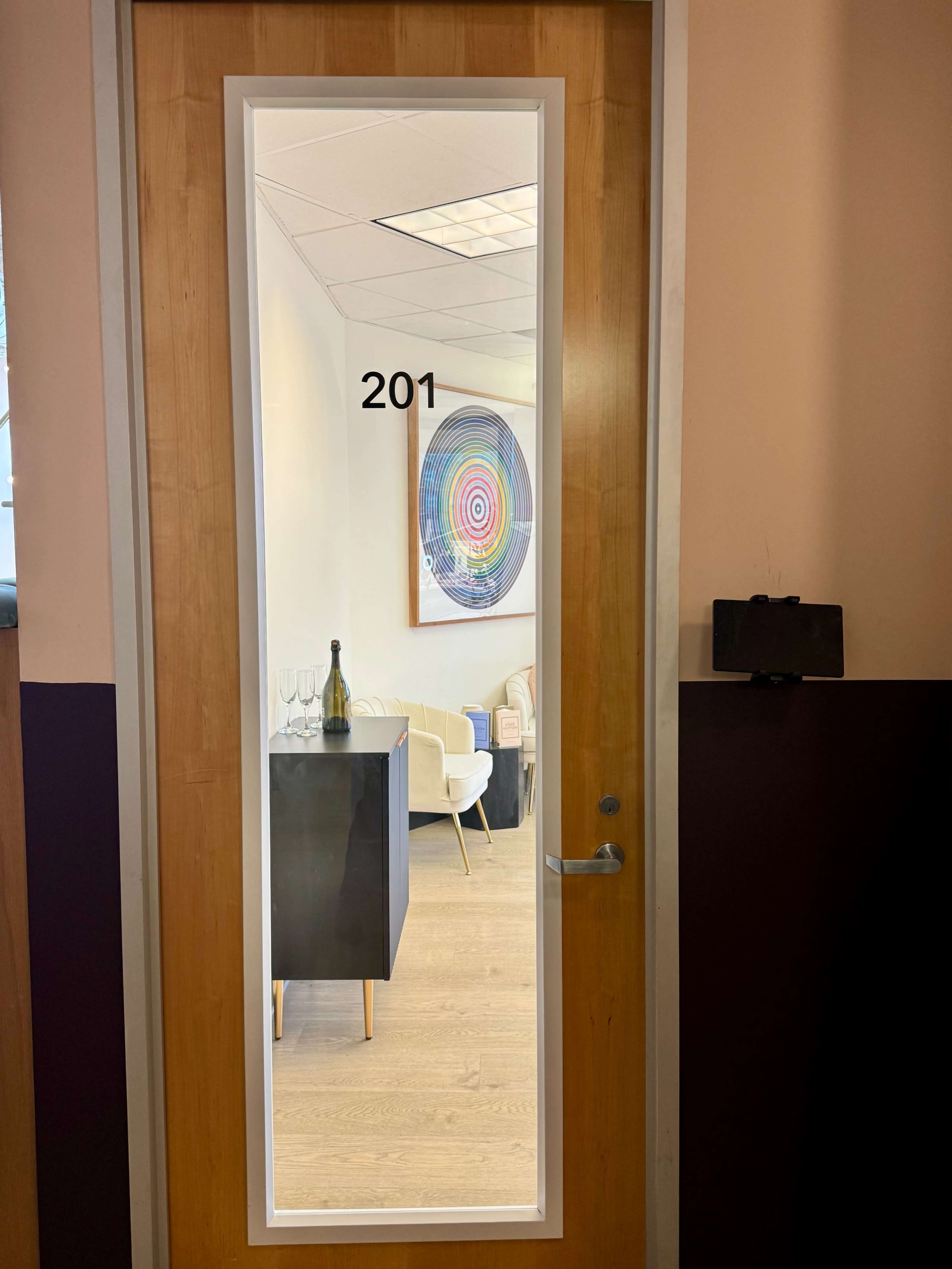 The image shows a wooden door marked "201," with a glass panel revealing an interior space featuring a colorful circular artwork and modern furniture.