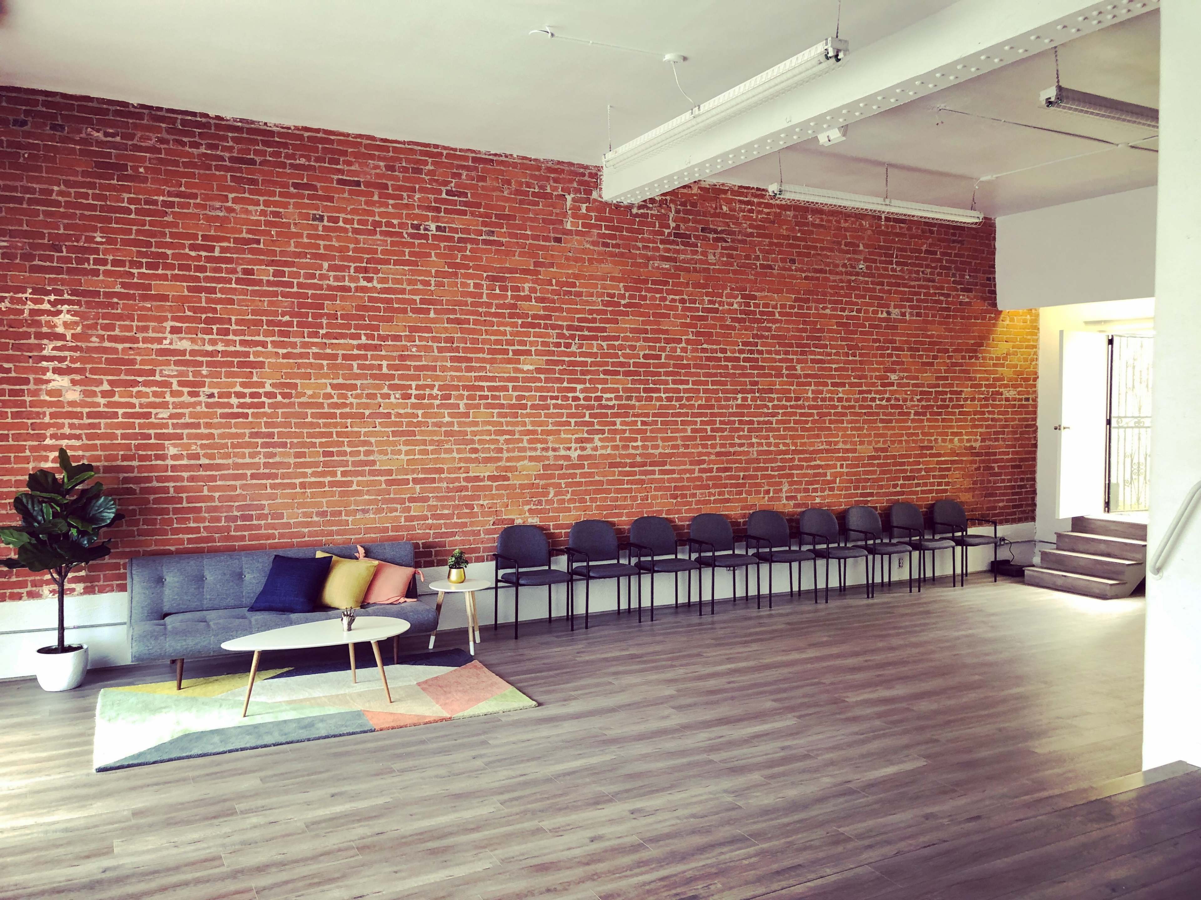 A spacious room with a brick wall, a gray sofa with colorful pillows, a patterned rug, and a row of chairs along one side.