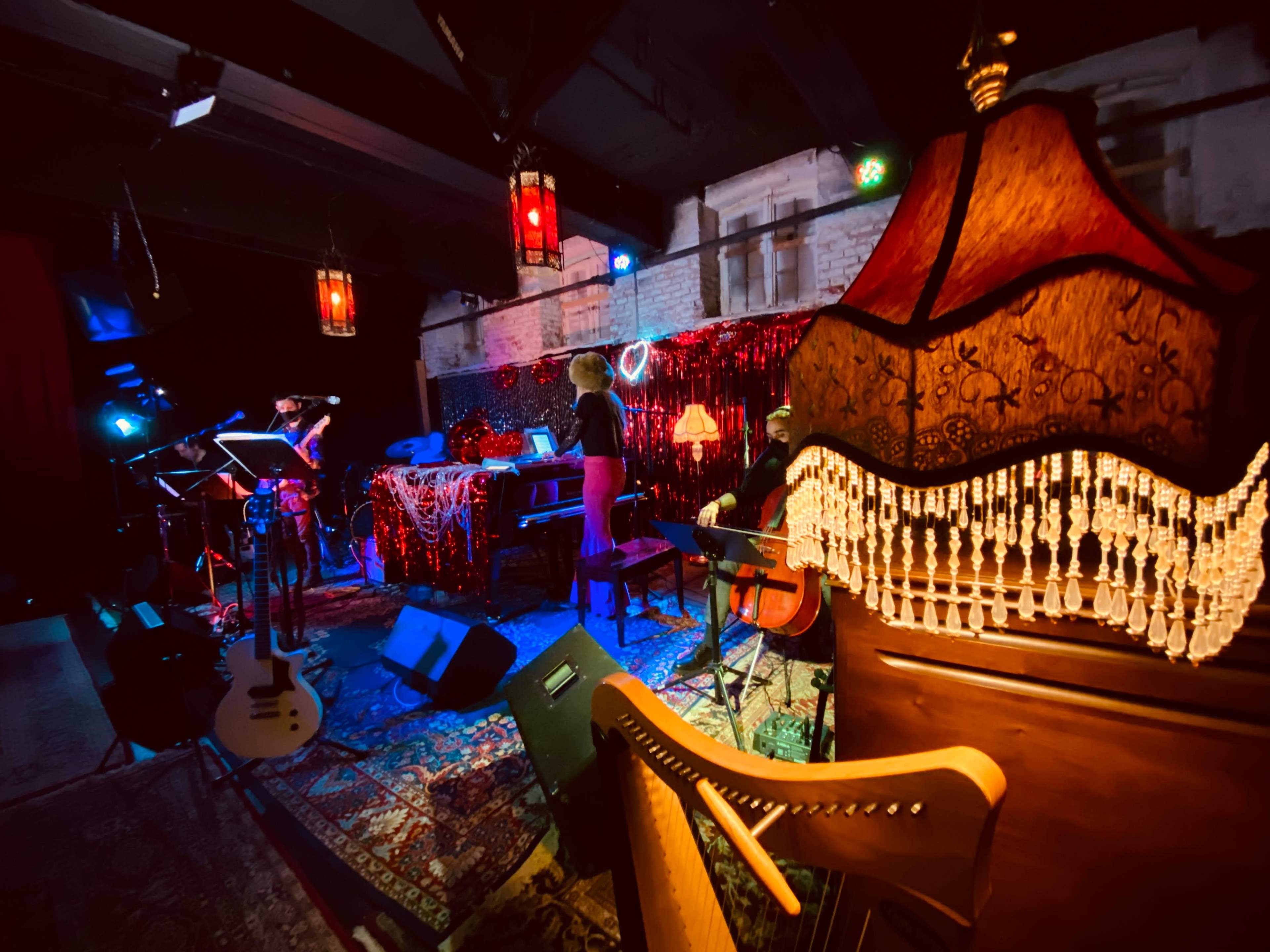 The image shows a dimly lit music venue with musicians on stage, surrounded by decorative elements like lamps and a harp.