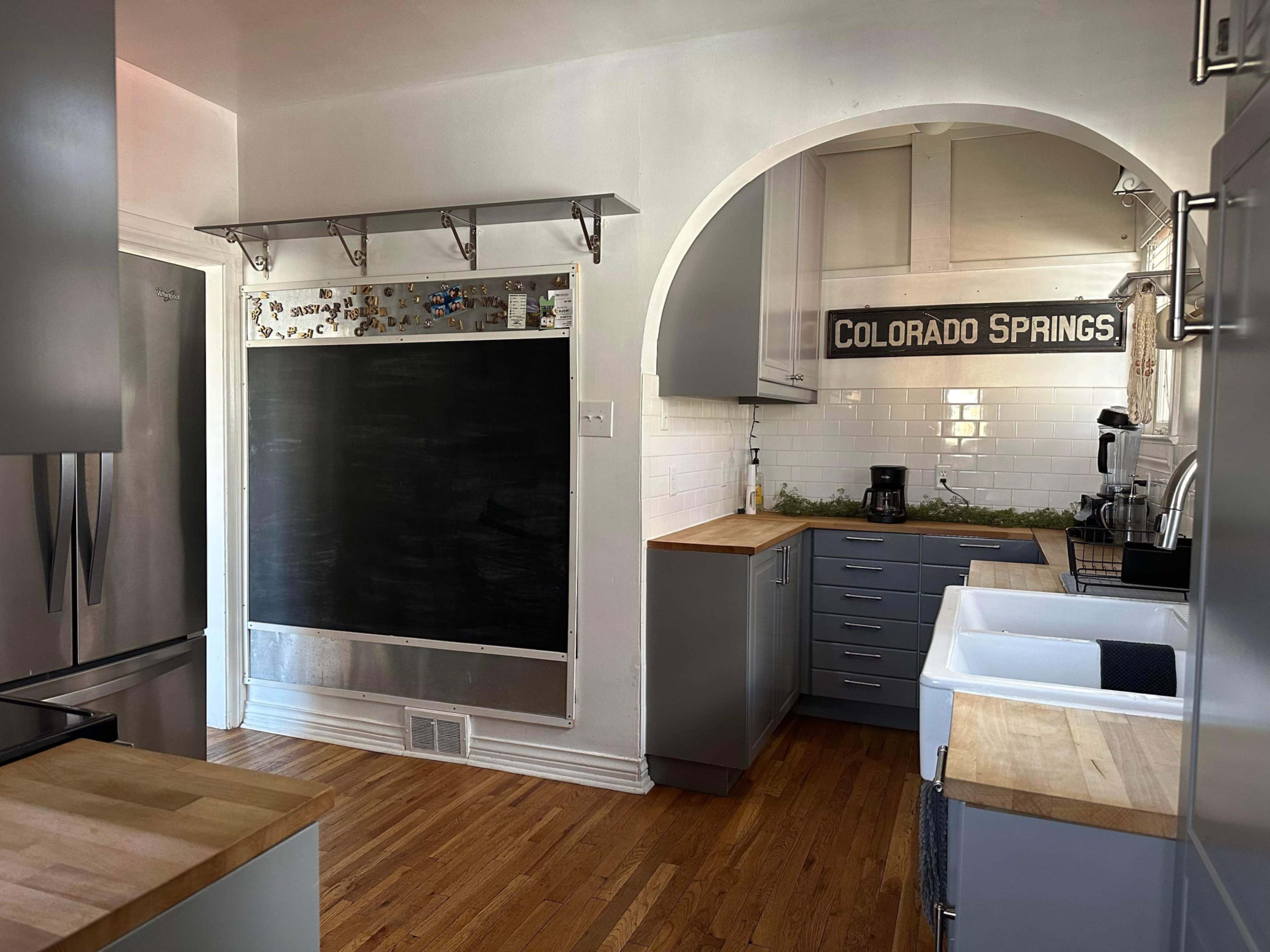 A modern kitchen with a large chalkboard on the left, stainless steel appliances, wooden countertops, and a sink beneath an arched entryway to another room.