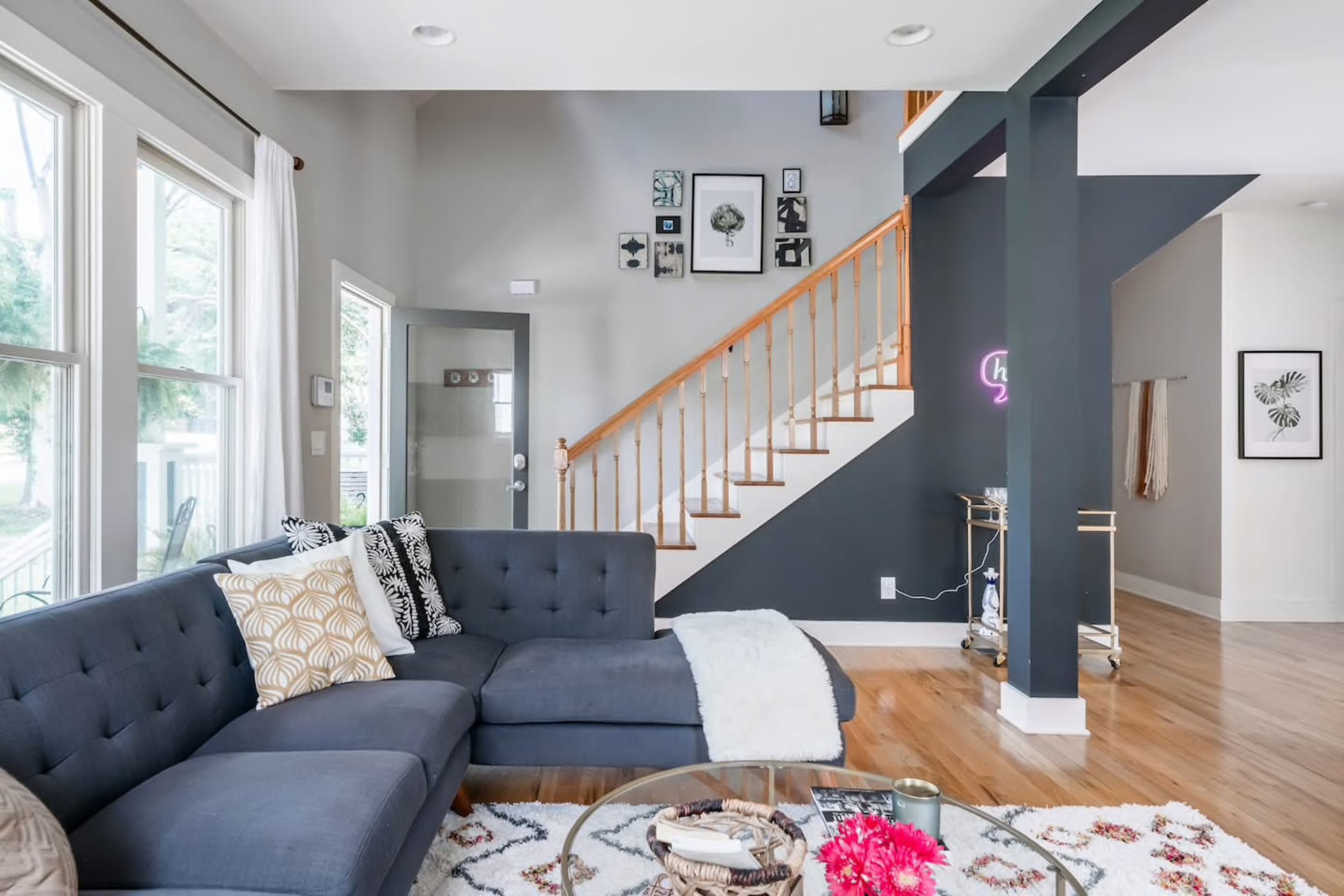 A modern living room features a sectional couch, a round glass coffee table, and a staircase adorned with framed artwork.