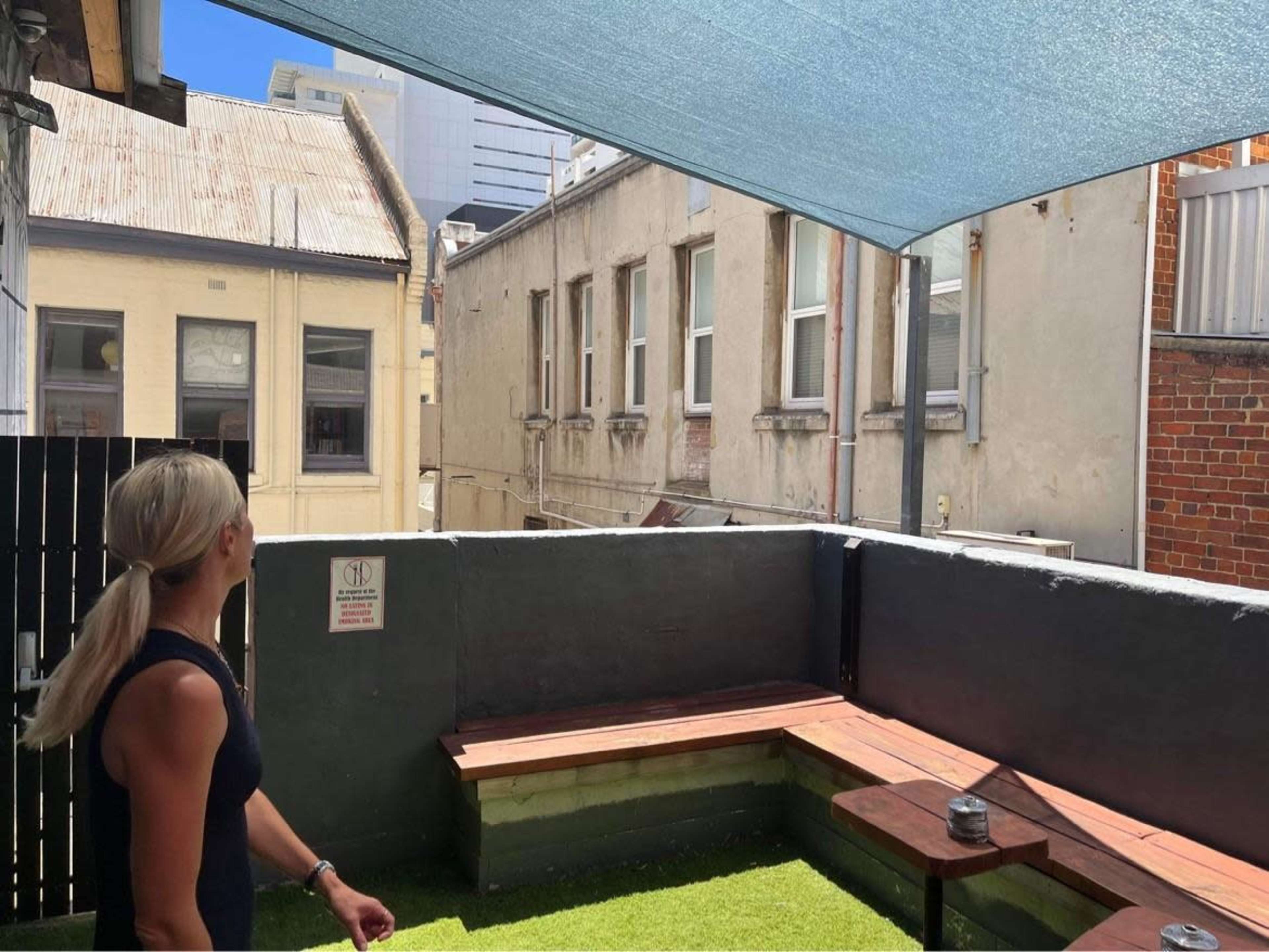 A woman stands on a rooftop terrace overlooking brick buildings and a blue sky, with a shaded area and seating made of wooden benches.
