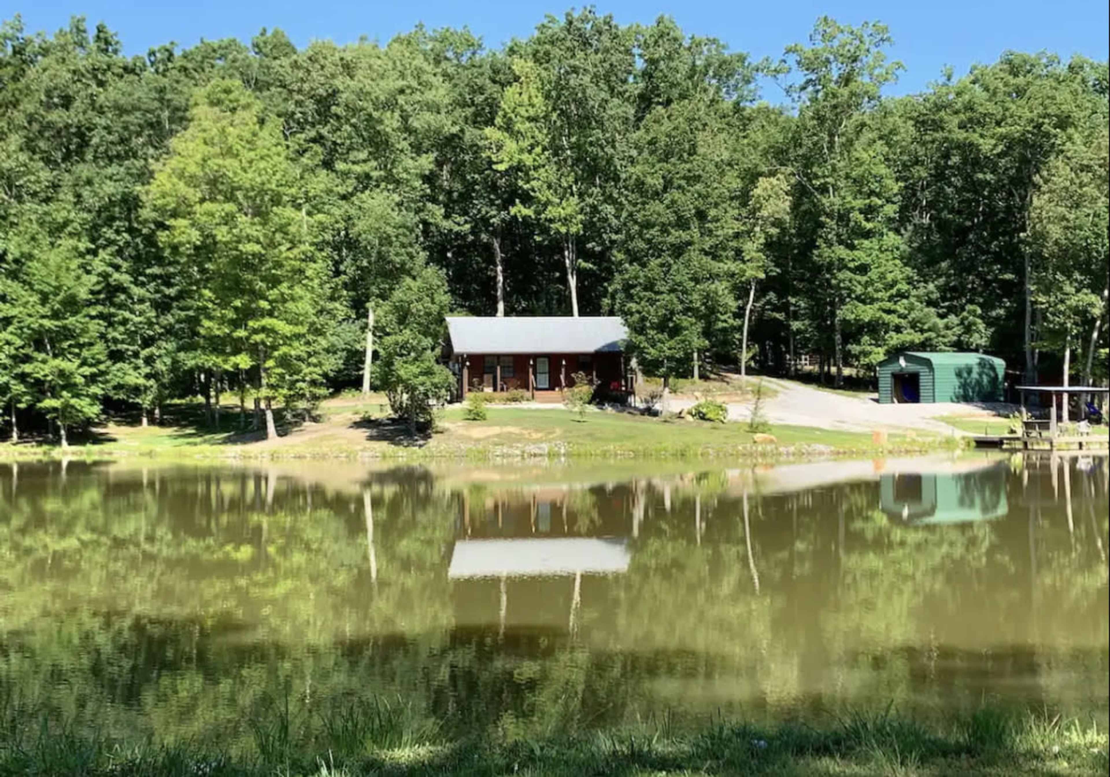 A wooden cabin with a gray roof is reflected in a calm pond surrounded by trees.