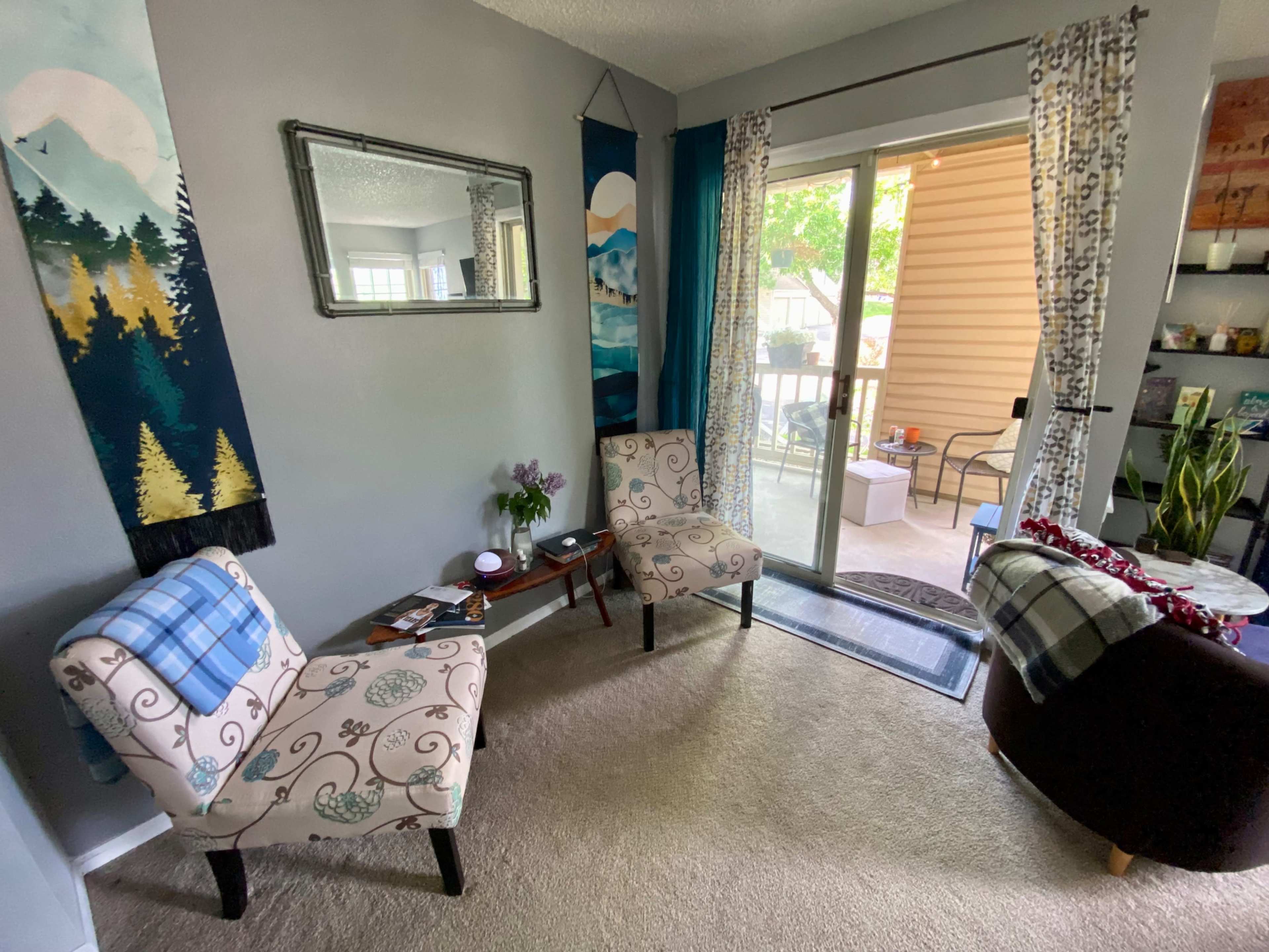 The image shows a cozy living room corner with two patterned chairs, a small table, and a sliding glass door leading to a patio.