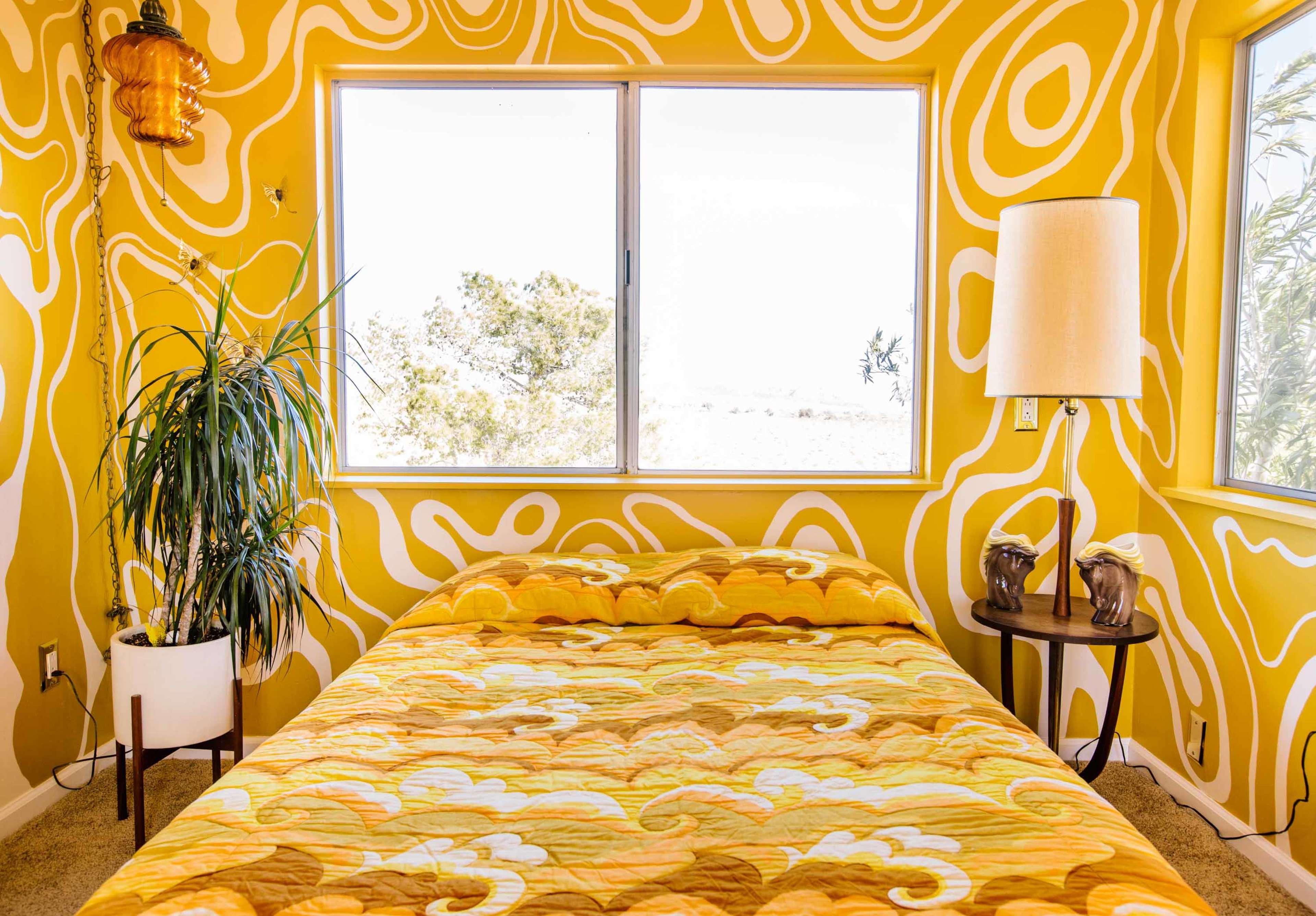 A bright yellow bedroom features a patterned wall, a bed with an orange and yellow quilt, a potted plant, and a lamp beside the bed.