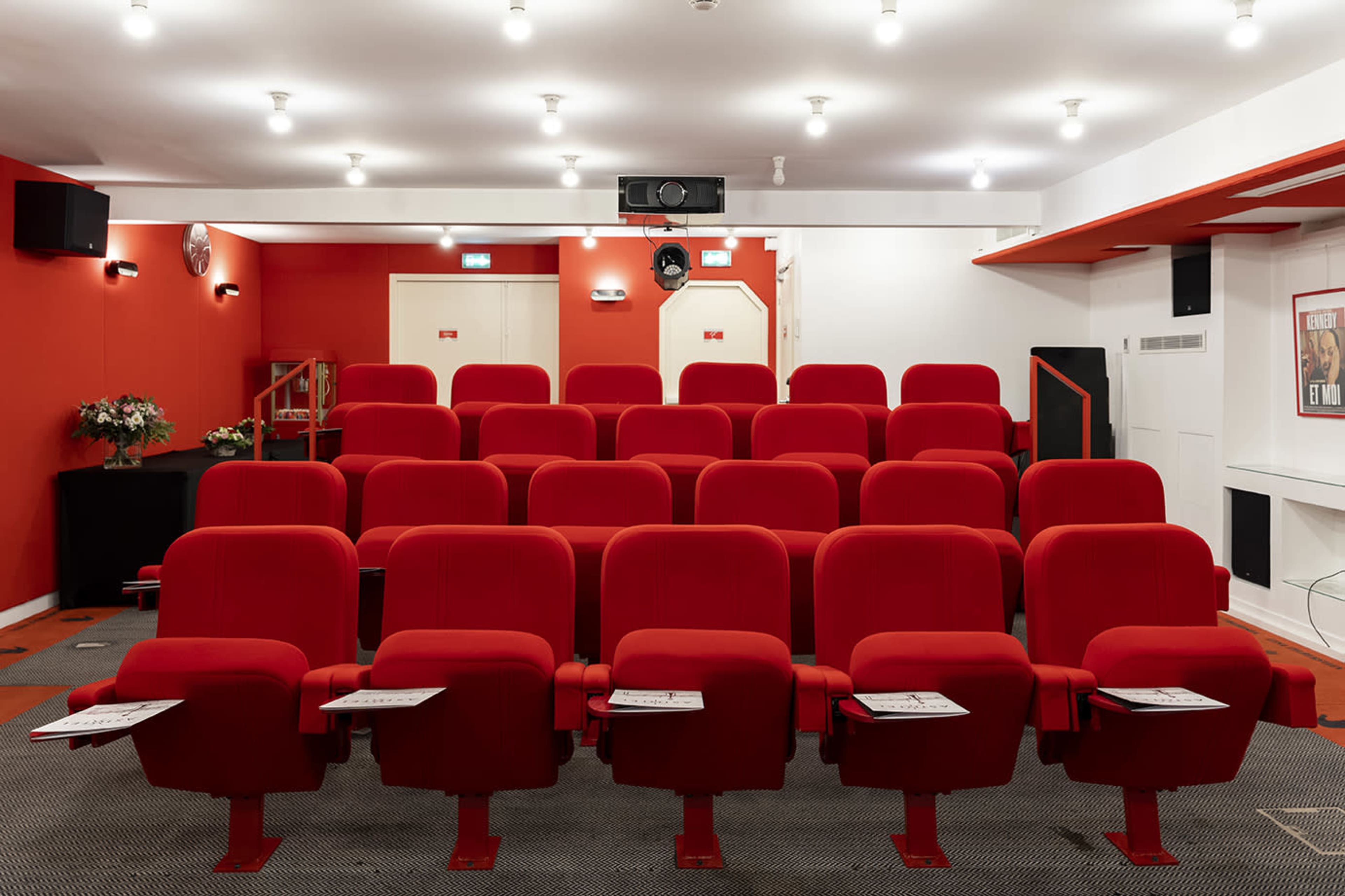 A small theater features red upholstered seats arranged in rows facing a projector screen, with a simple interior design.