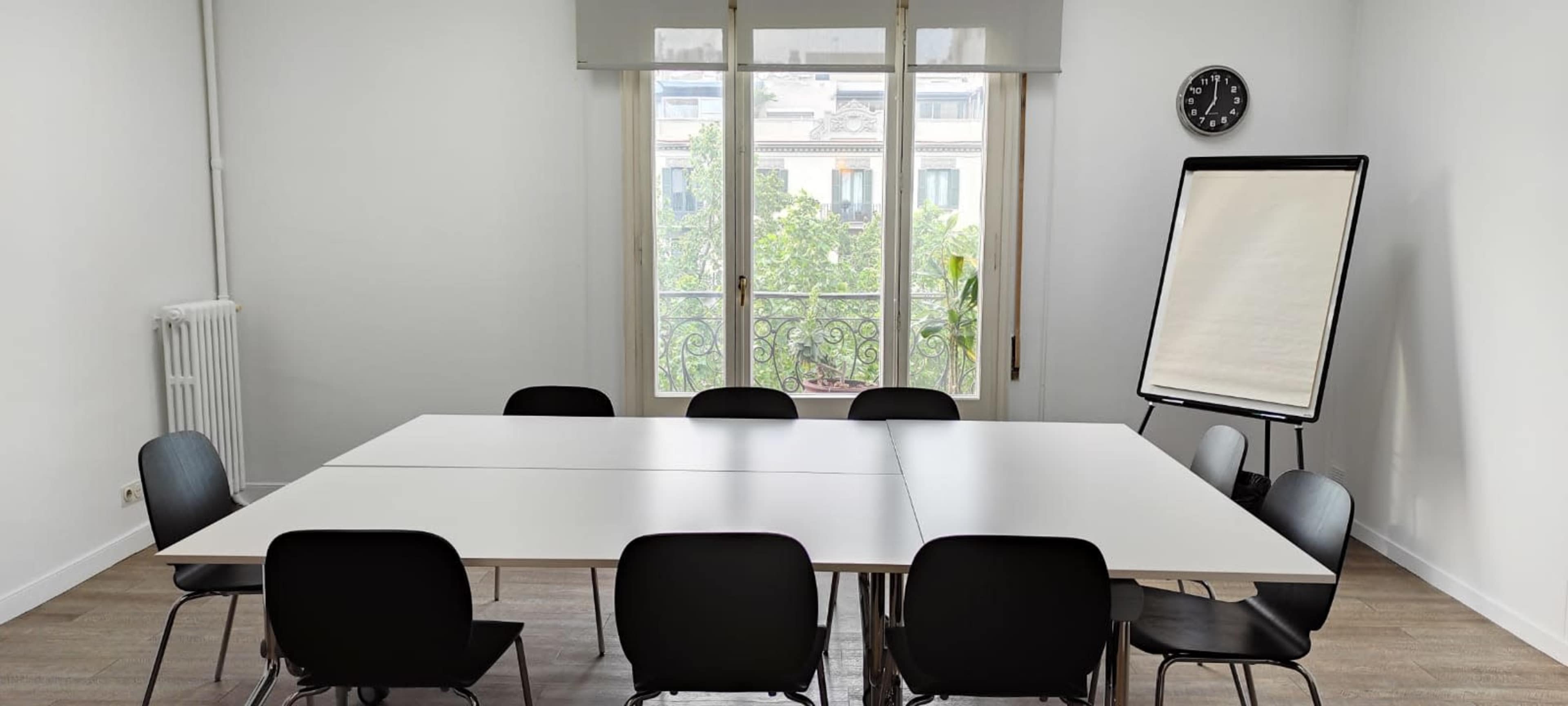 The image shows a modern meeting room with a large table surrounded by black chairs, a flip chart, a clock on the wall, and a window overlooking a green view.